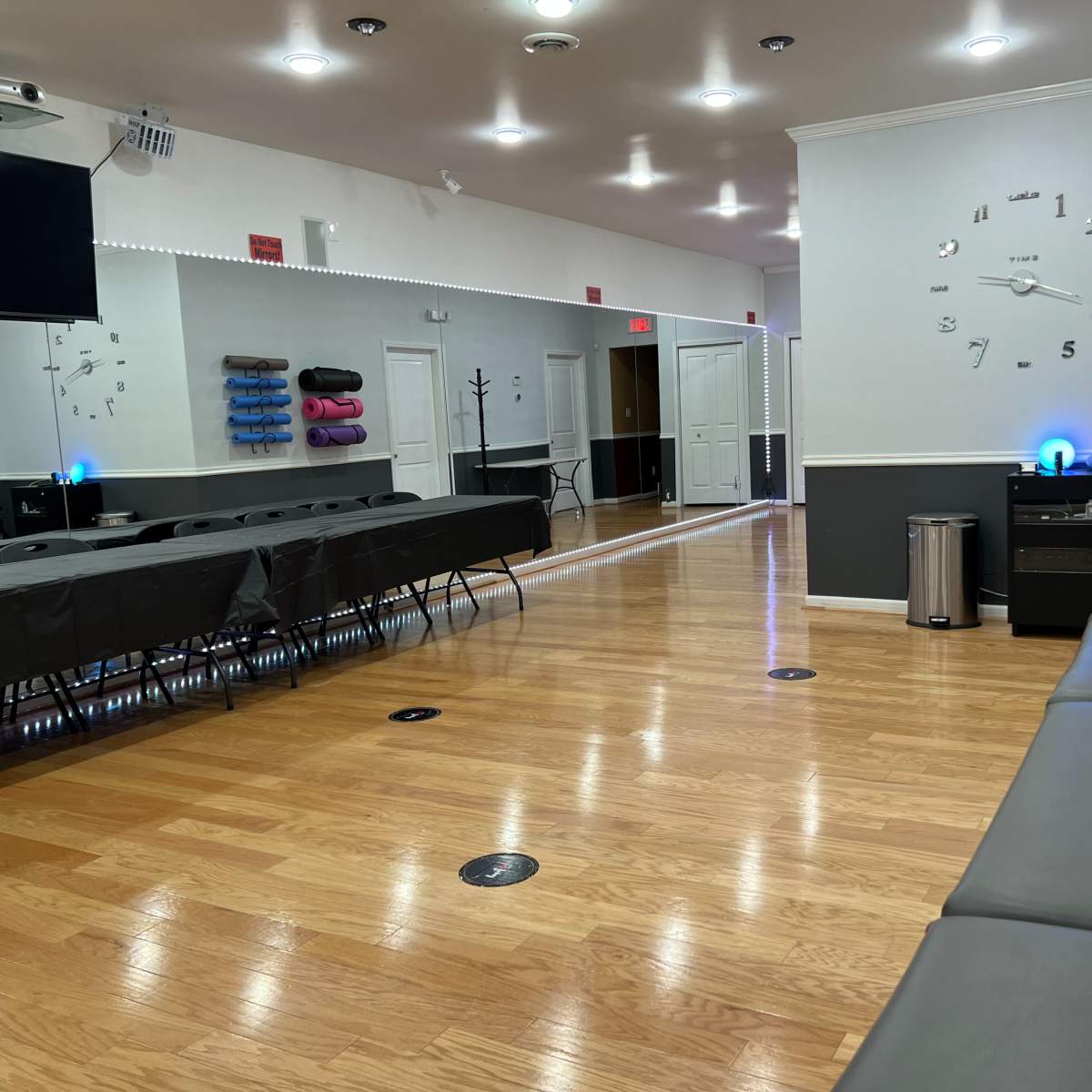 A brightly lit interior space with a row of black tables and chairs, a wall clock, and a long bench against the wall.