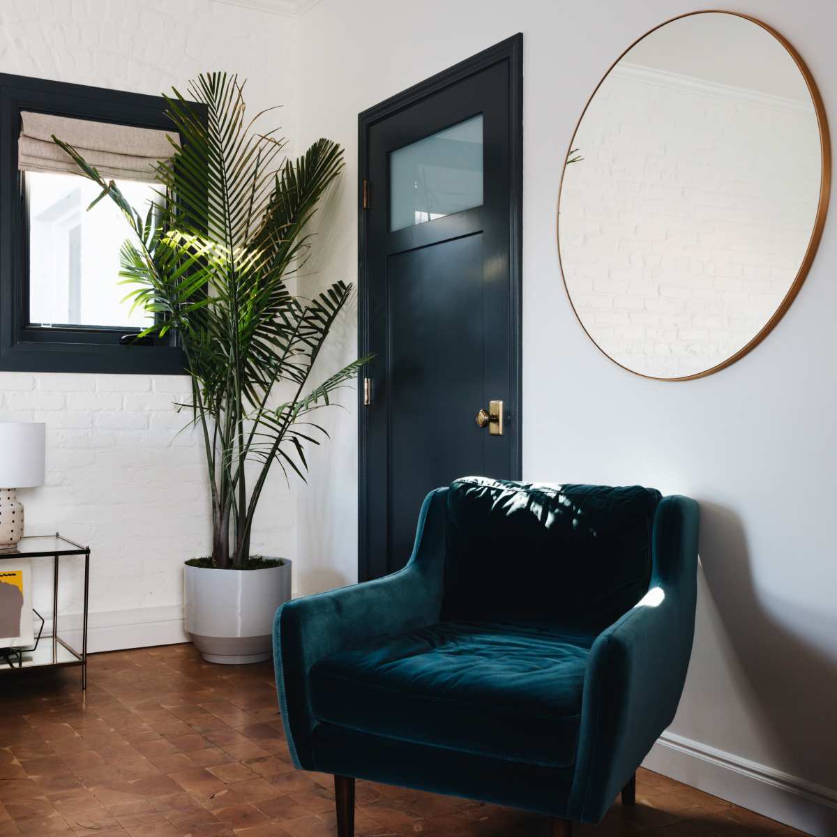 A teal velvet armchair is positioned next to a tall indoor plant, with a round mirror and a white lamp on a small table in a well-lit room.
