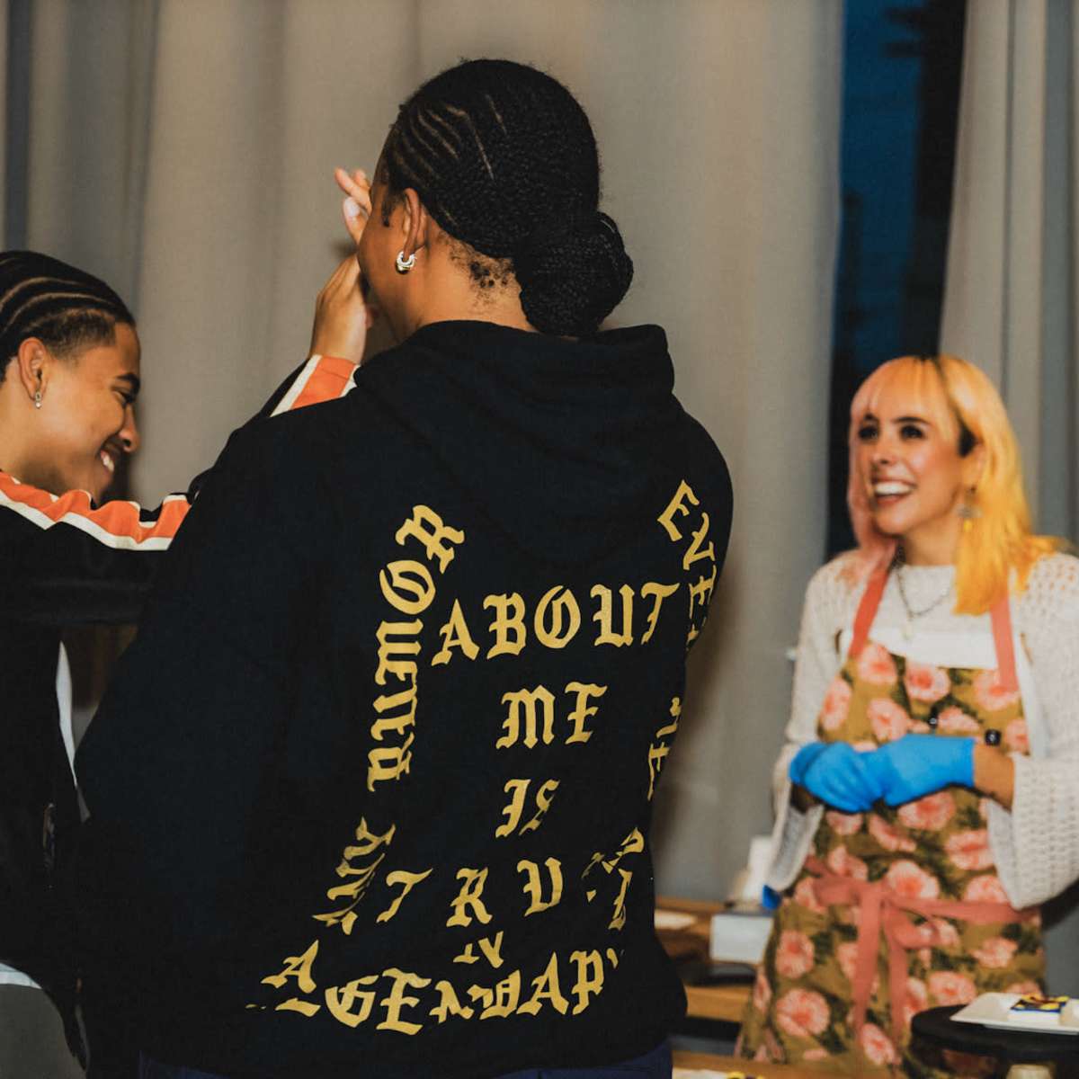 A group of people interacts in a dining setting, with one person wearing an apron and blue gloves, while two others engage in conversation.