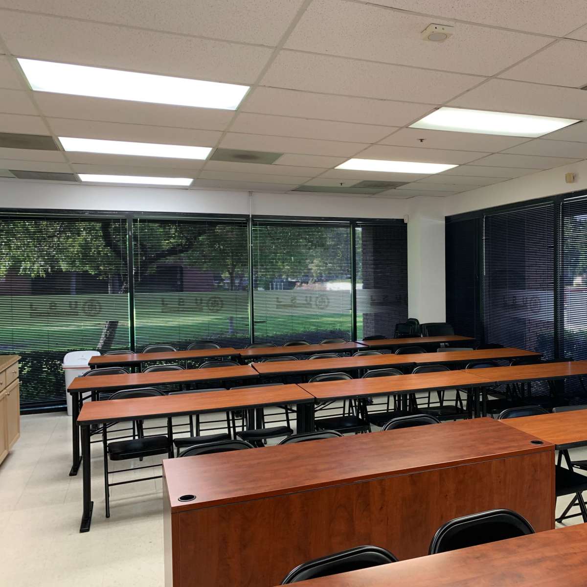 A classroom with several rows of wooden tables and black chairs, illuminated by overhead lights and large windows covered by blinds.