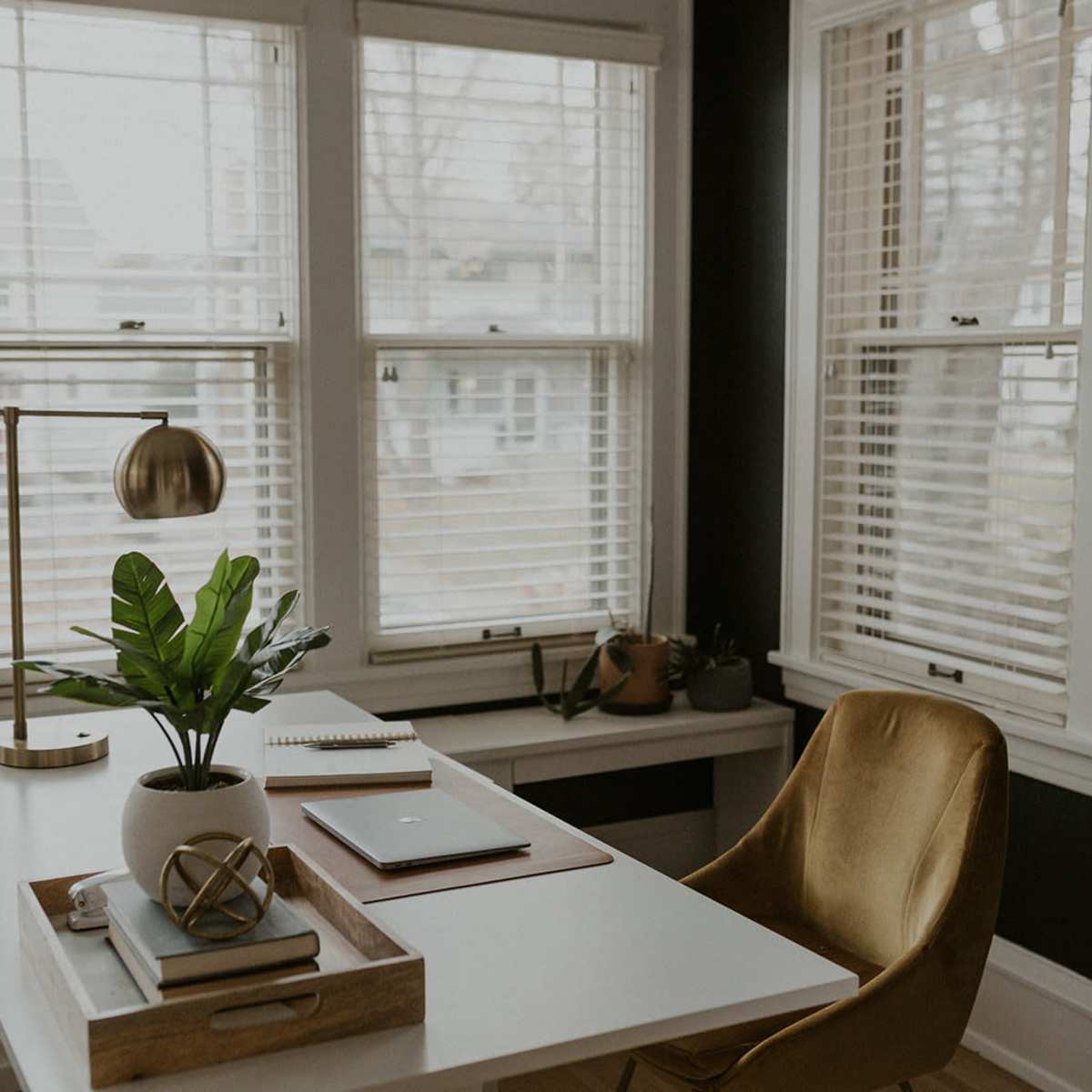 A modern office space features a white desk with a laptop, notebook, and decorative items, accompanied by a gold desk lamp and a green plant, set against large windows with blinds.