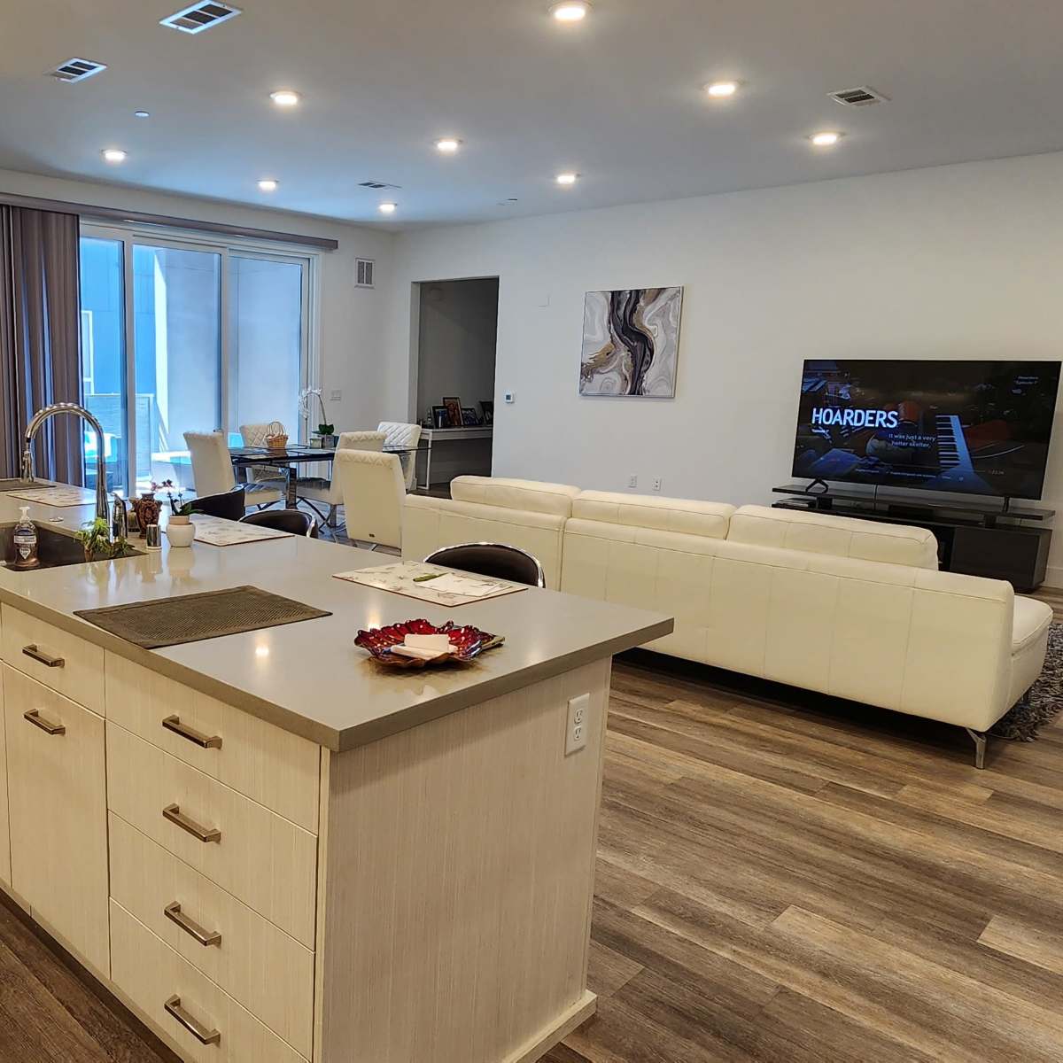 A modern kitchen and living area feature light-colored cabinetry, a central island, and a television mounted on the wall in an open-concept space.
