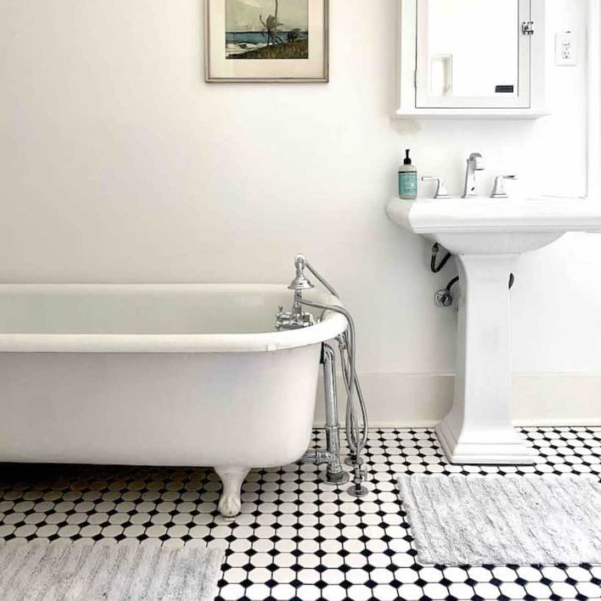 A bathroom with a freestanding white bathtub, a pedestal sink, and a wall-mounted mirror, all set against a backdrop of white walls and black and white tiled floor.