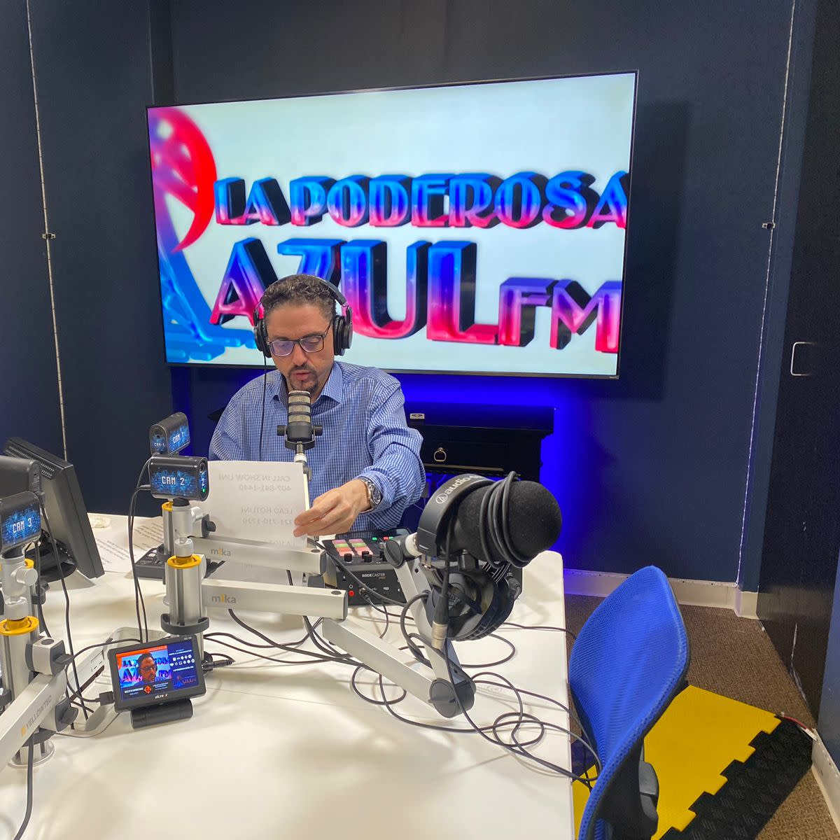 A man wearing headphones reads from a paper while seated at a radio broadcast table with microphones and screens, in front of a large display that says "LA PODEROSA AZUL FM."