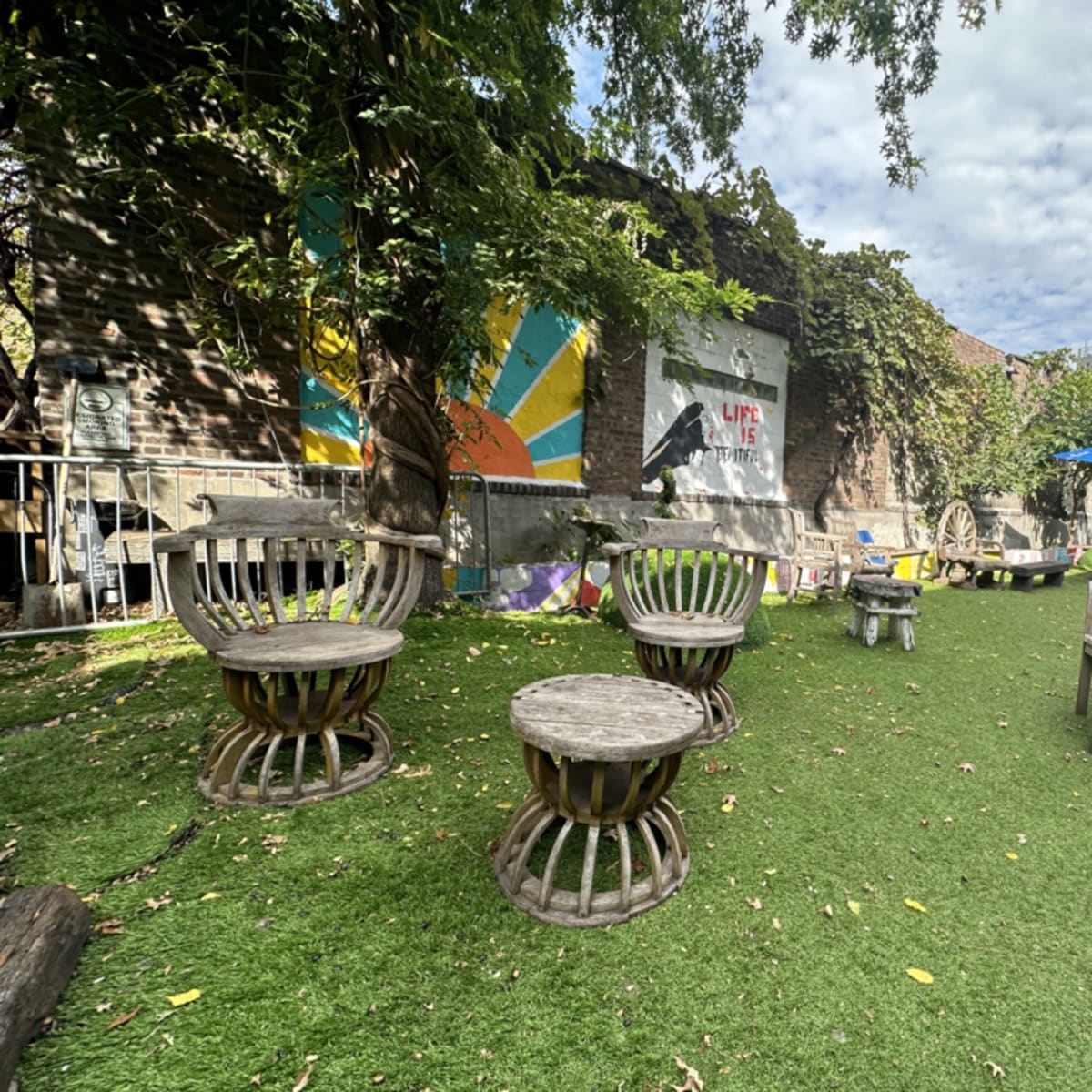 An outdoor seating area with wooden chairs and tables set on artificial grass surrounded by greenery and a colorful mural on the wall.