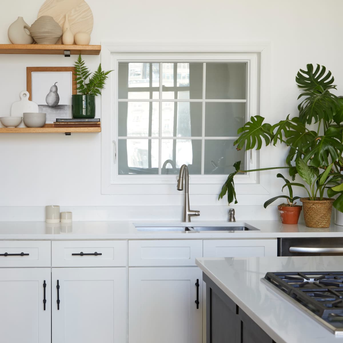 A modern kitchen features white cabinetry, open wooden shelves with decorative items, a stainless steel sink, and a large window.