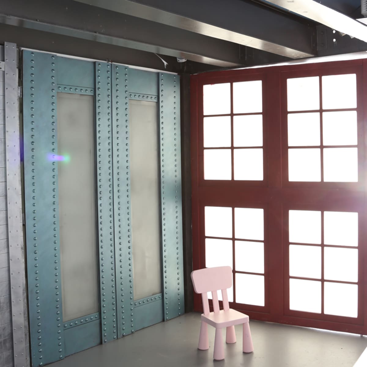 A pink wooden chair stands alone in a well-lit room with large windows and metallic blue walls.