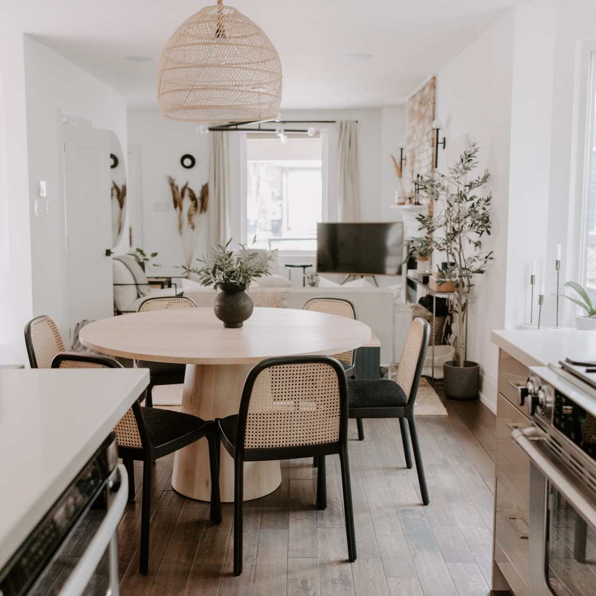 A dining area features a round wooden table surrounded by four chairs, with a modern kitchen and living space visible in the background.