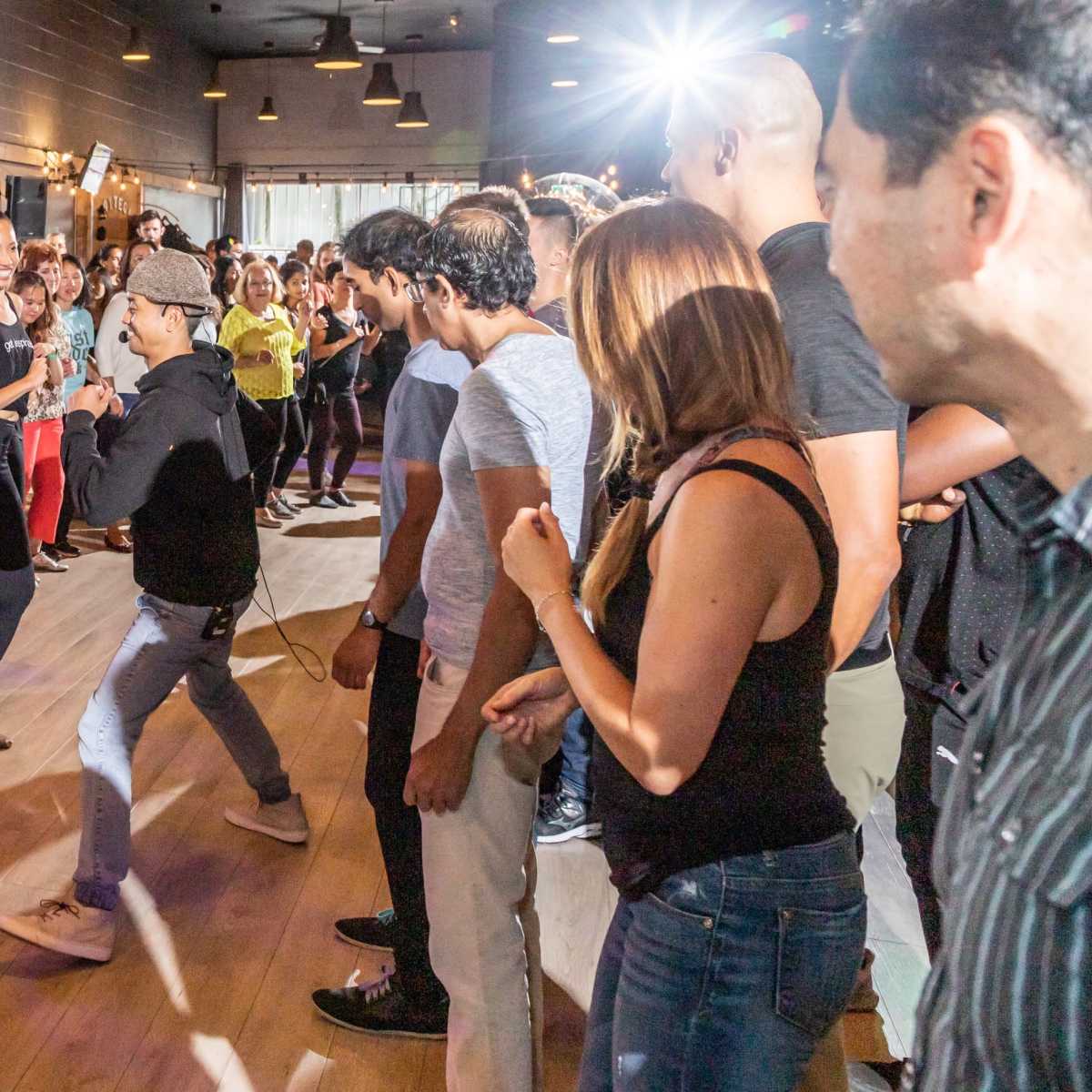 A large group of people dance in a lively studio with lights and a wooden floor.