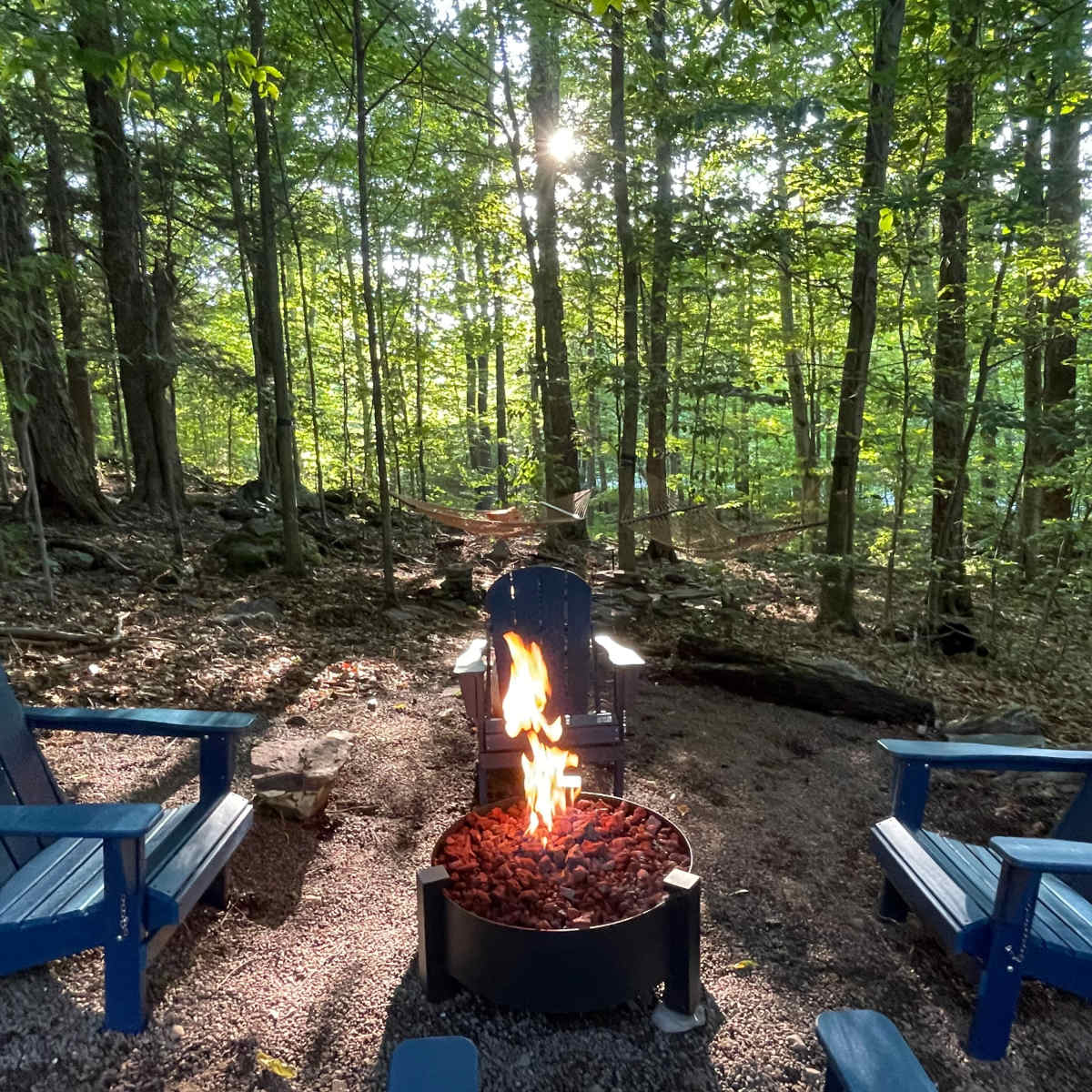 A circular fire pit with a flame surrounded by blue adirondack chairs sits in a forested area with trees and a hammock in the background.