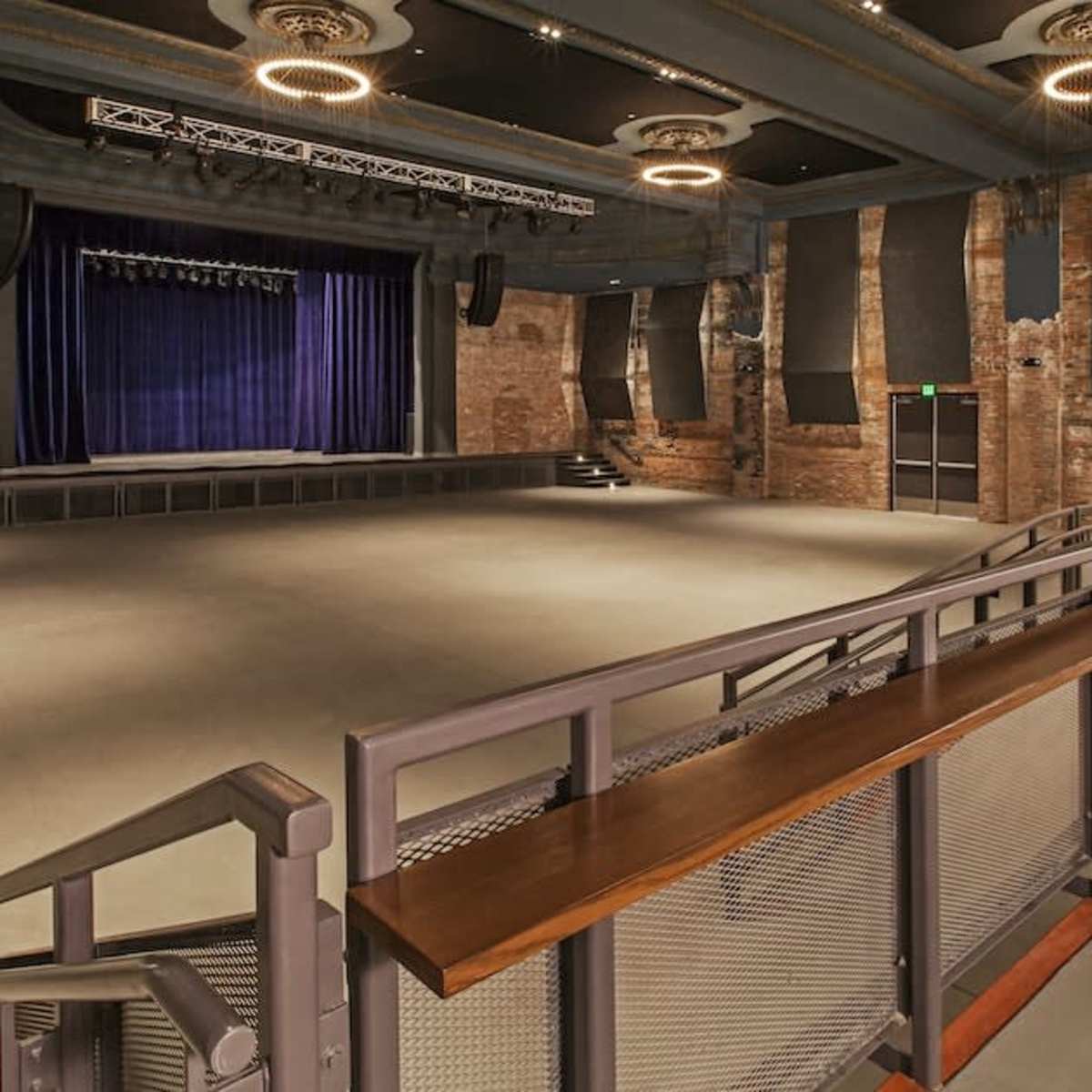An empty auditorium with a wooden railing overlooking a stage surrounded by exposed brick walls and spotlights.