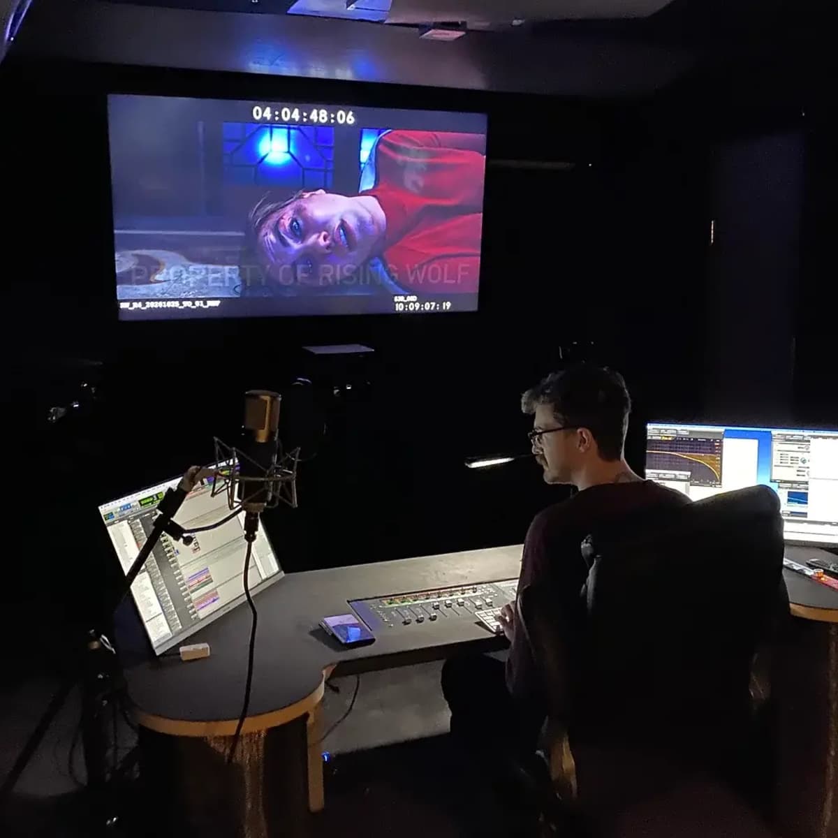 A person sits at a control desk in a dark studio, monitoring a screen displaying a scene from a film.