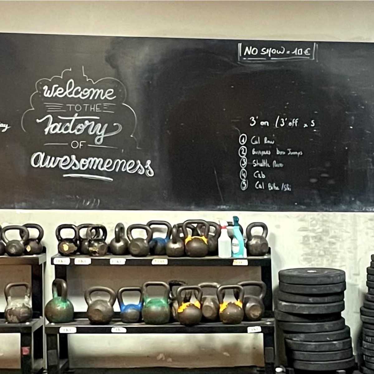 A gym space with a chalkboard displaying a welcome message and gym rules, alongside rows of kettlebells and weight plates on shelves.