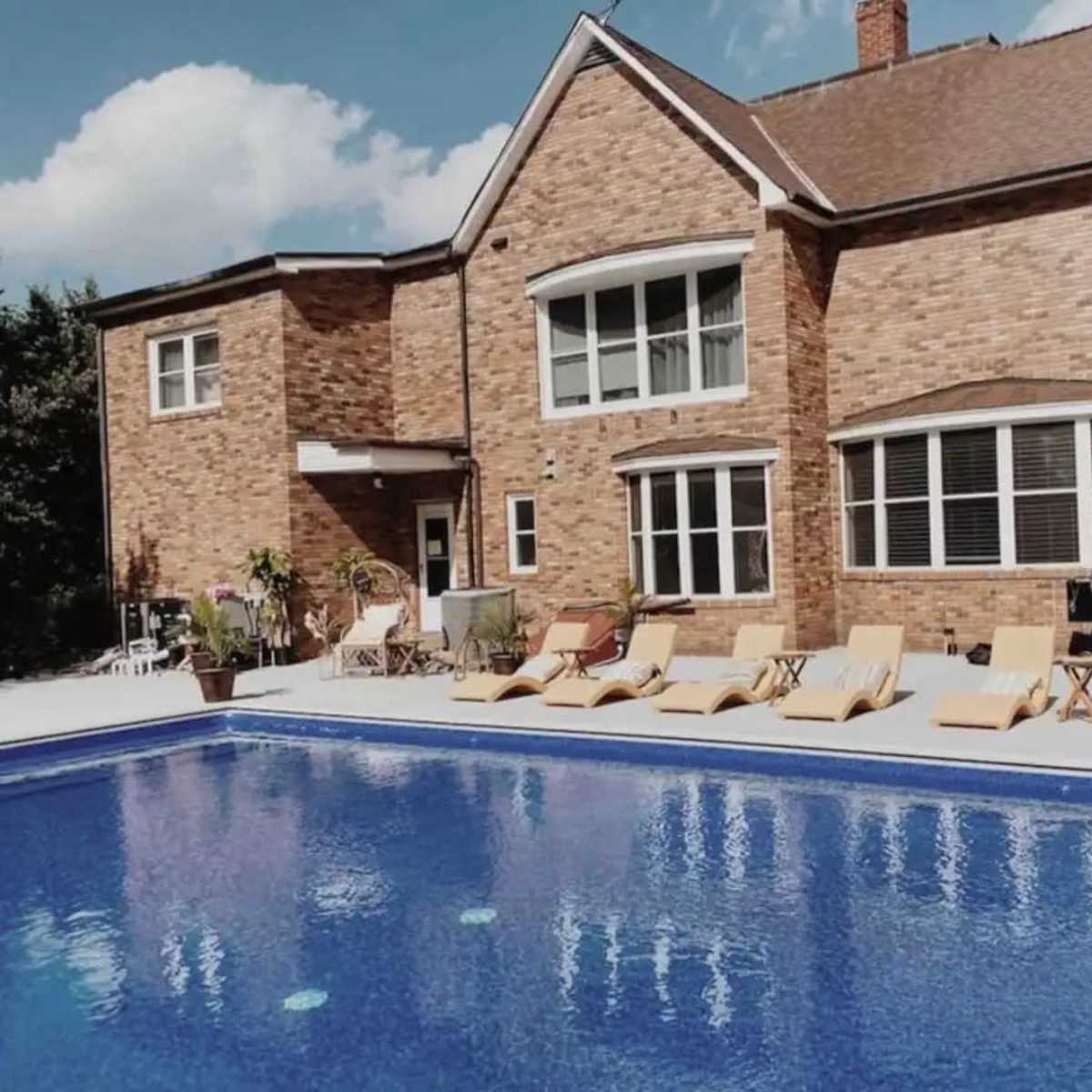 A spacious brick house with a large swimming pool surrounded by lounge chairs and potted plants.