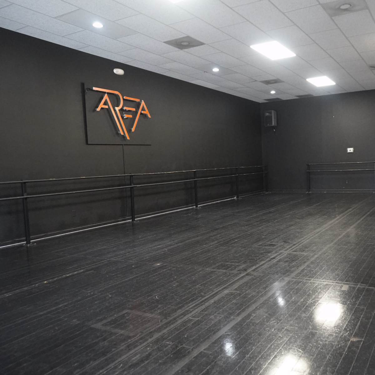 A dance studio featuring a black wall with a logo, wooden flooring, and ballet bars along one side.