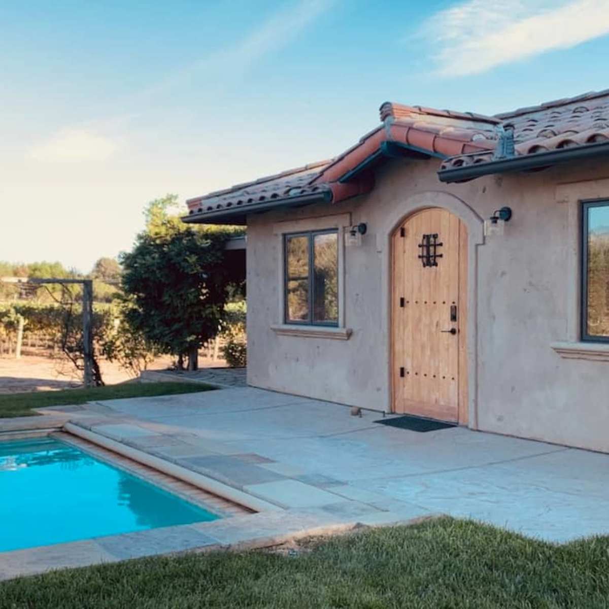 A house with a tiled roof beside a swimming pool, surrounded by a grassy area and vineyards in the background.