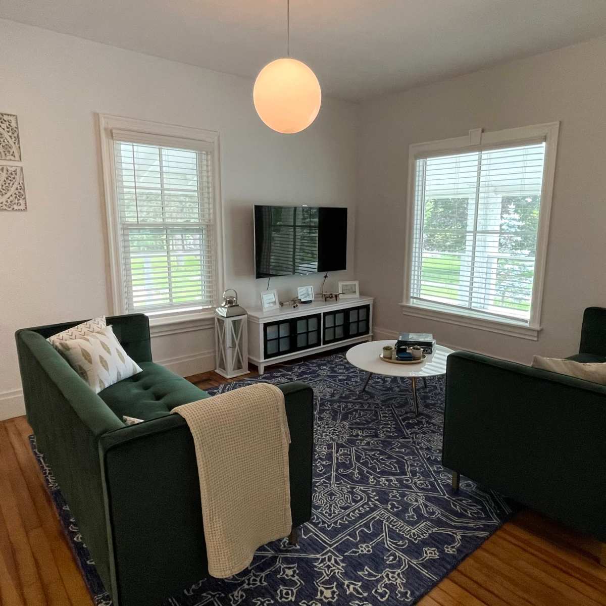 A cozy living room with two green sofas facing a coffee table, a television mounted on the wall, and large windows letting in natural light.
