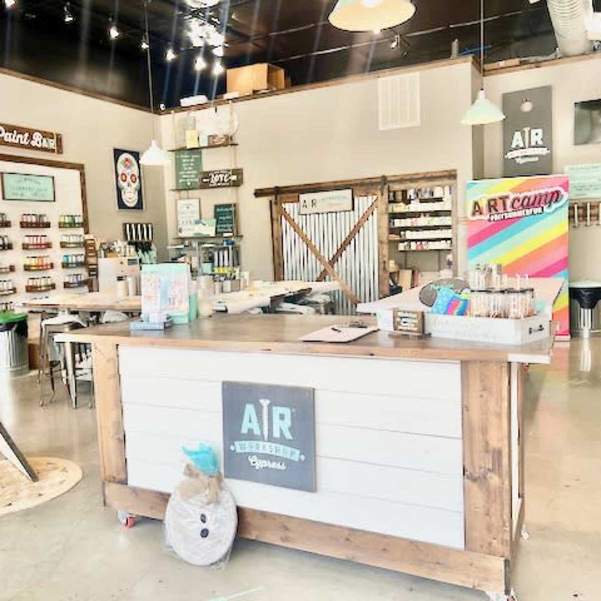 An art supply store with a central service counter, shelves filled with paint, and a colorful promotional display for an art camp.