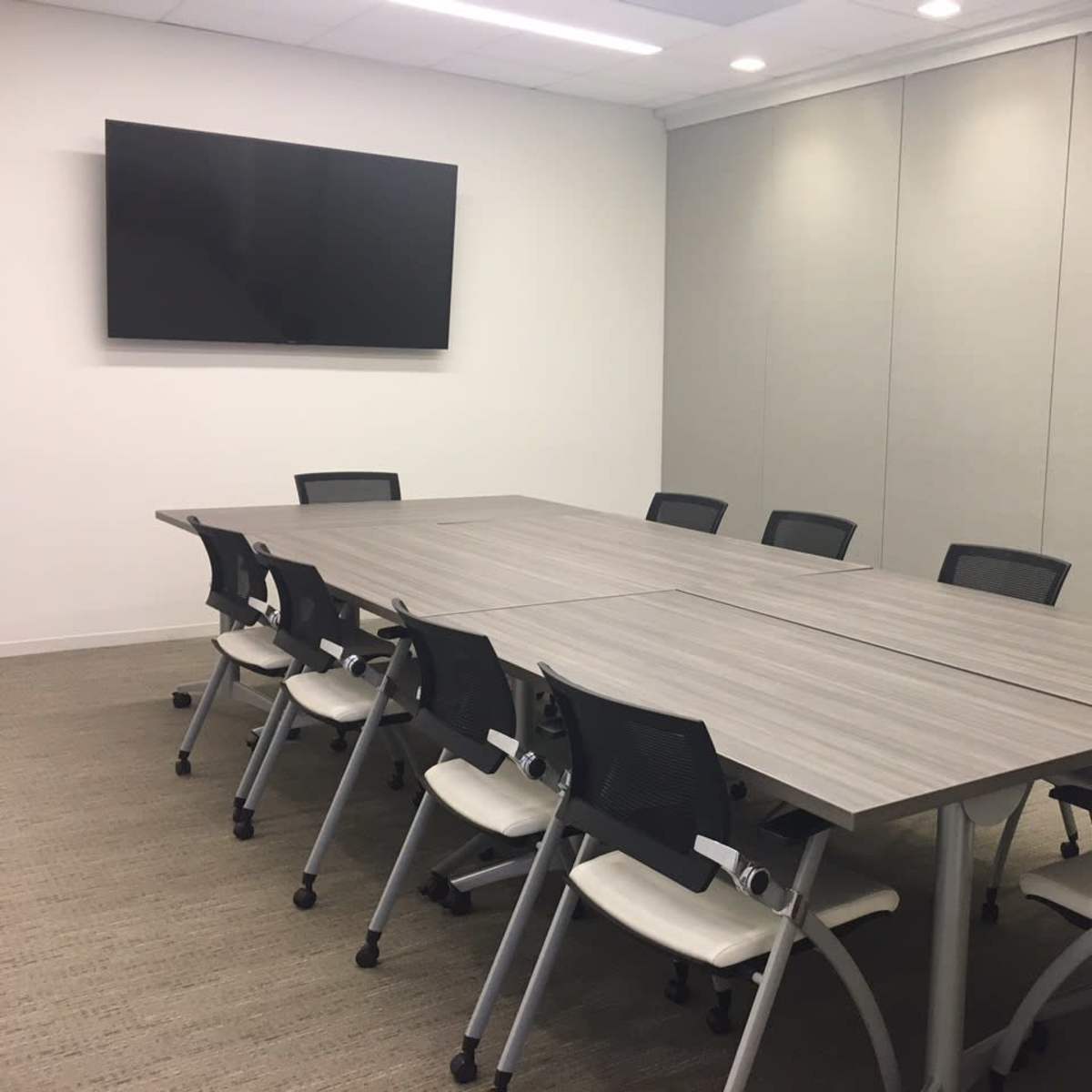 A spacious conference room features a large, rectangular table surrounded by eight chairs, with a wall-mounted television in the background.