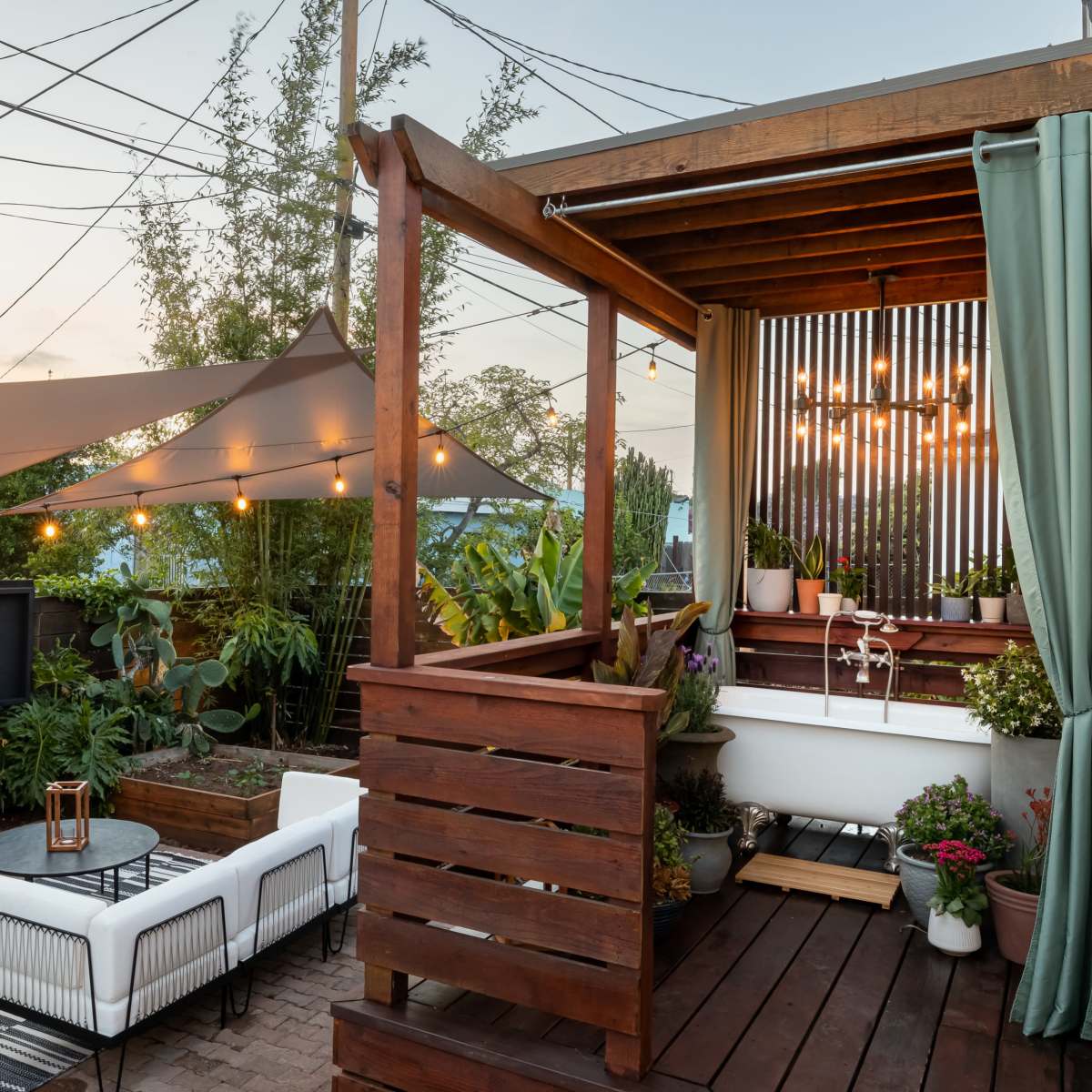 A backyard with a wooden deck, outdoor seating, and a large television, surrounded by plants and illuminated by string lights.