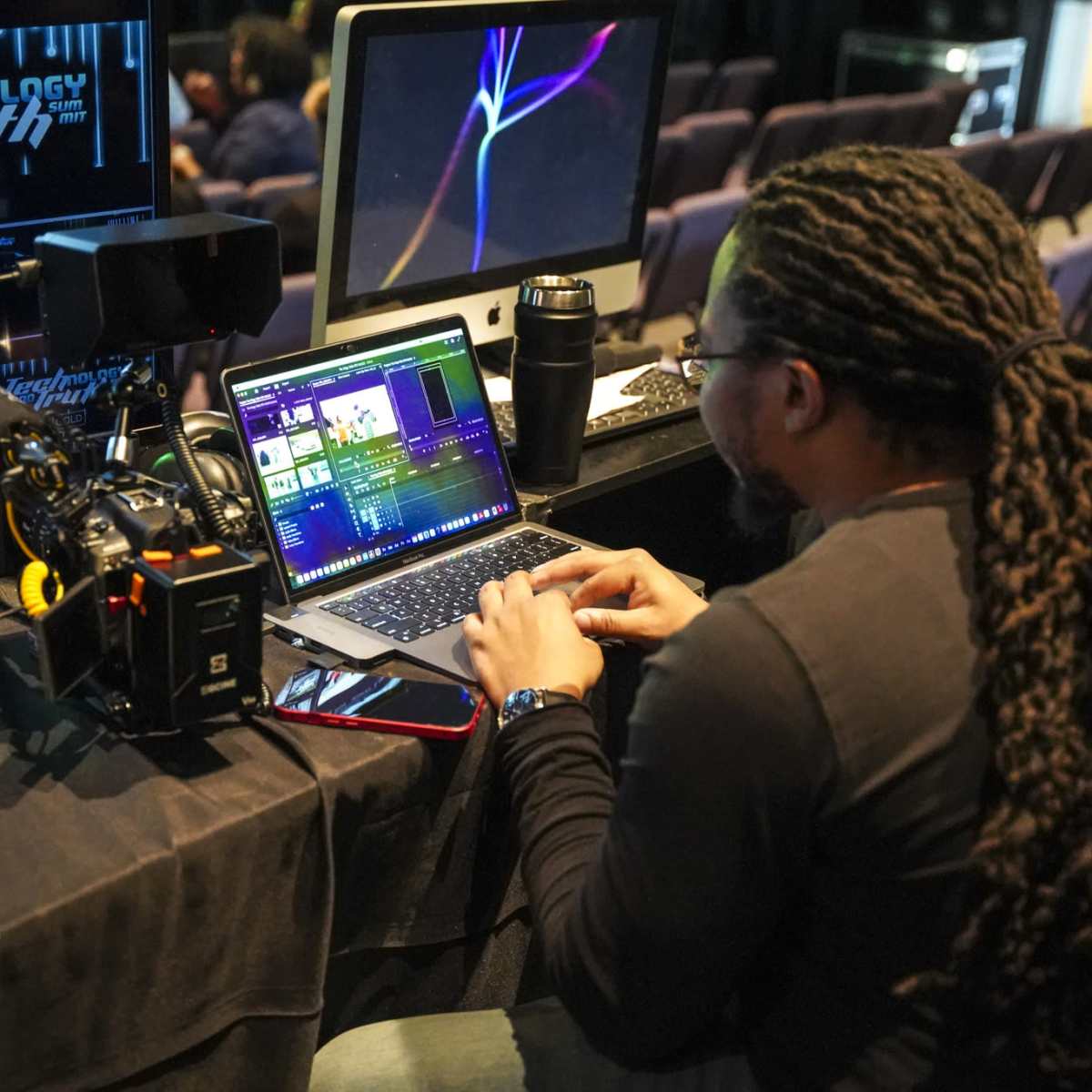A person with long hair works on video editing software on a laptop while seated at a desk with a camera and monitors displaying graphics.