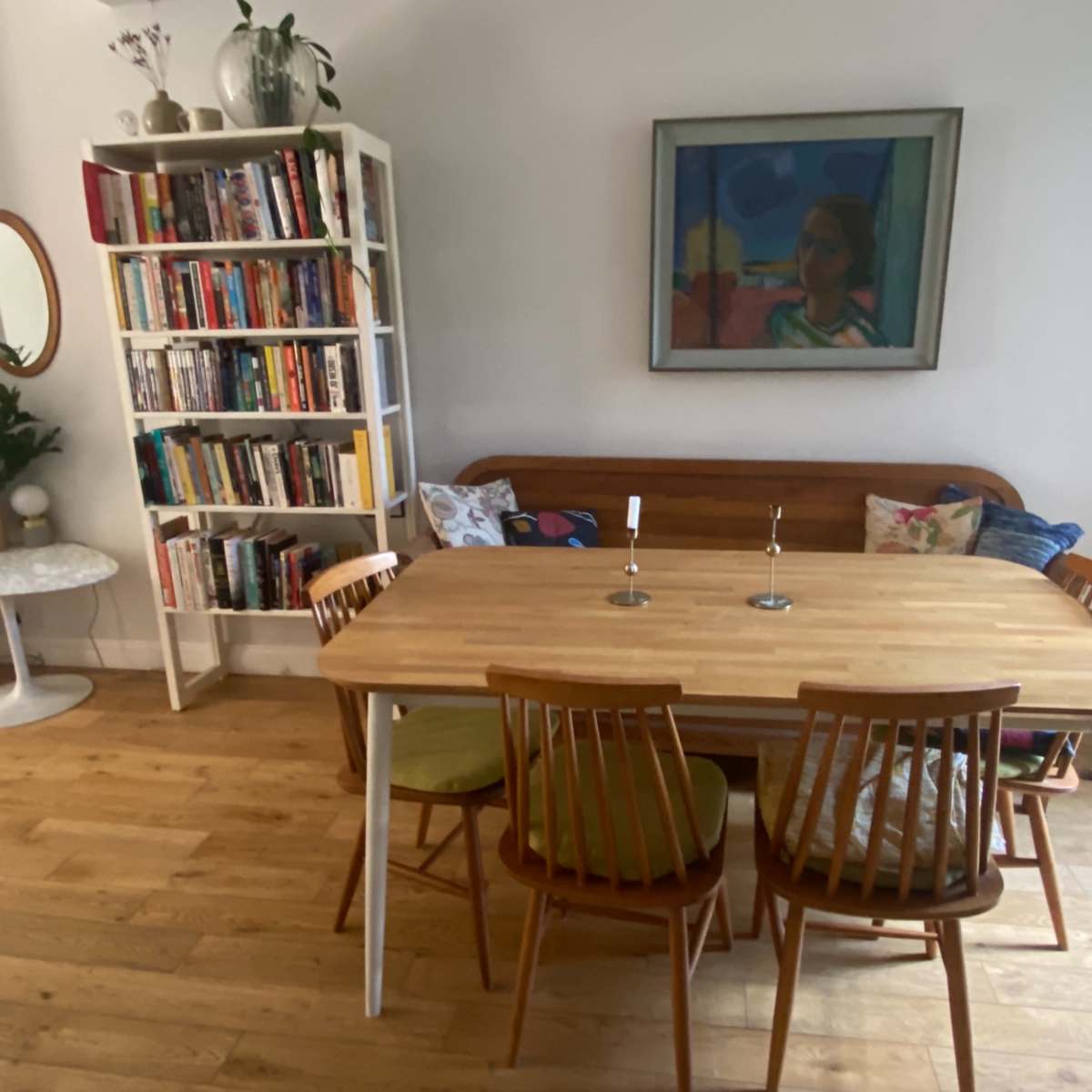 A dining area features a wooden table with four chairs, a bookshelf filled with books, and a colorful painting on the wall.