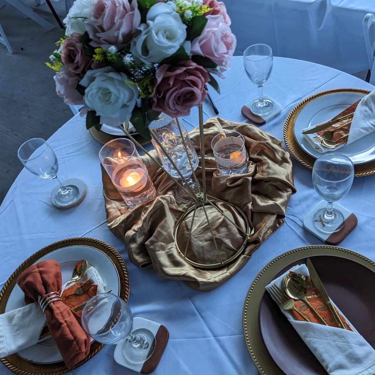 A round dining table is set with elegant plates, glassware, and a centerpiece of flowers, draped with a gold fabric.