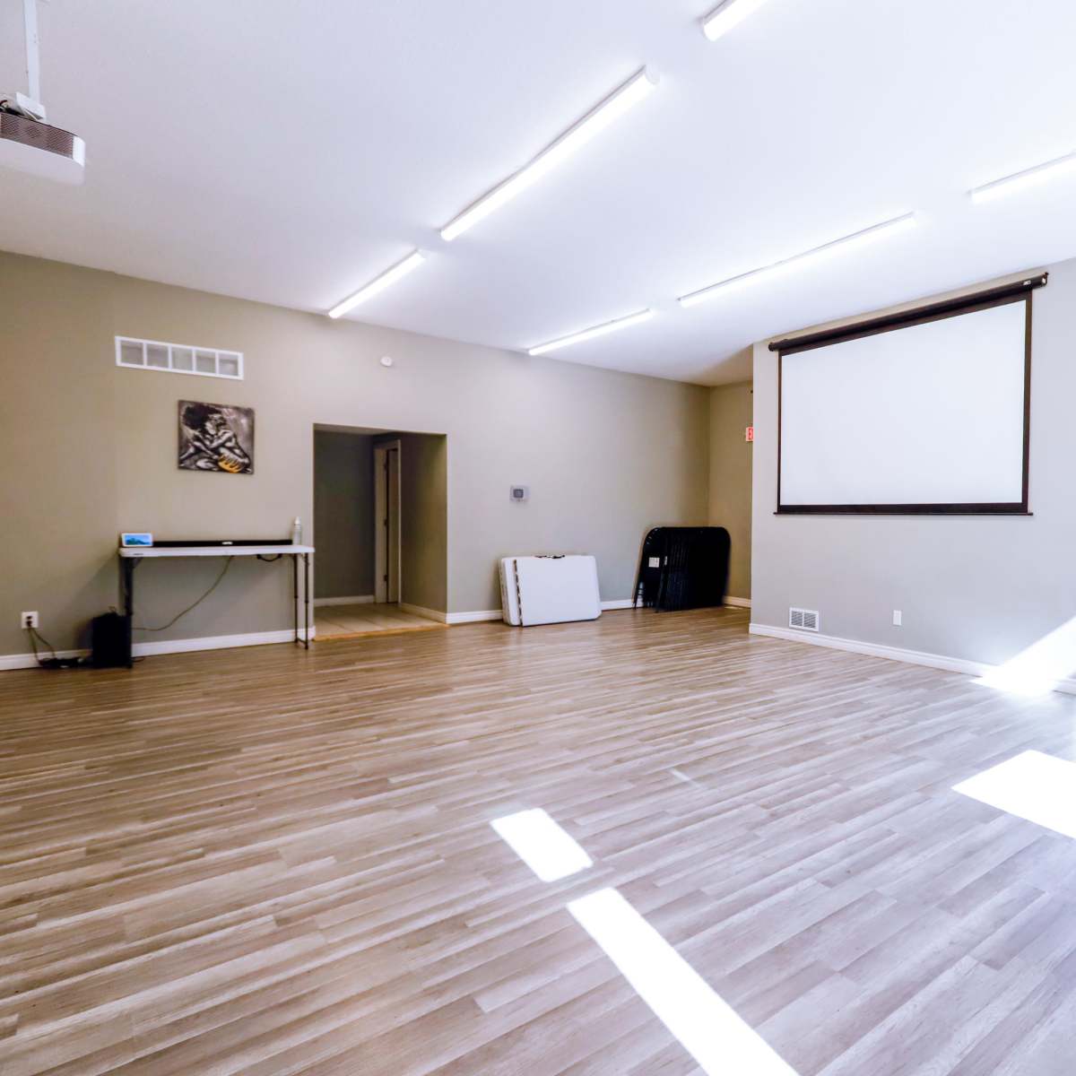 An empty room with wooden flooring, a large projection screen on one wall, and a set of folding tables stored against another wall.