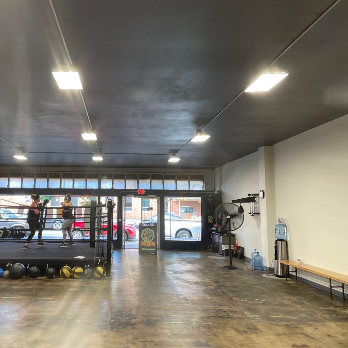 An indoor boxing gym with a boxing ring, training equipment, and large windows facing the street.