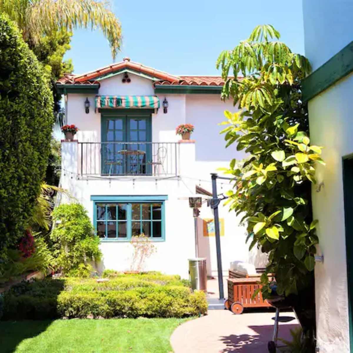 A narrow pathway leads to a two-story house surrounded by green hedges and a well-maintained garden with a patio.