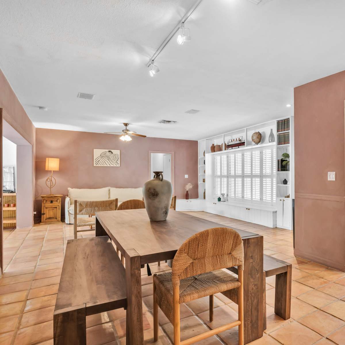 A spacious room with a dining table and chairs, a living area with a sofa and lamp, and a kitchen visible through an open doorway.