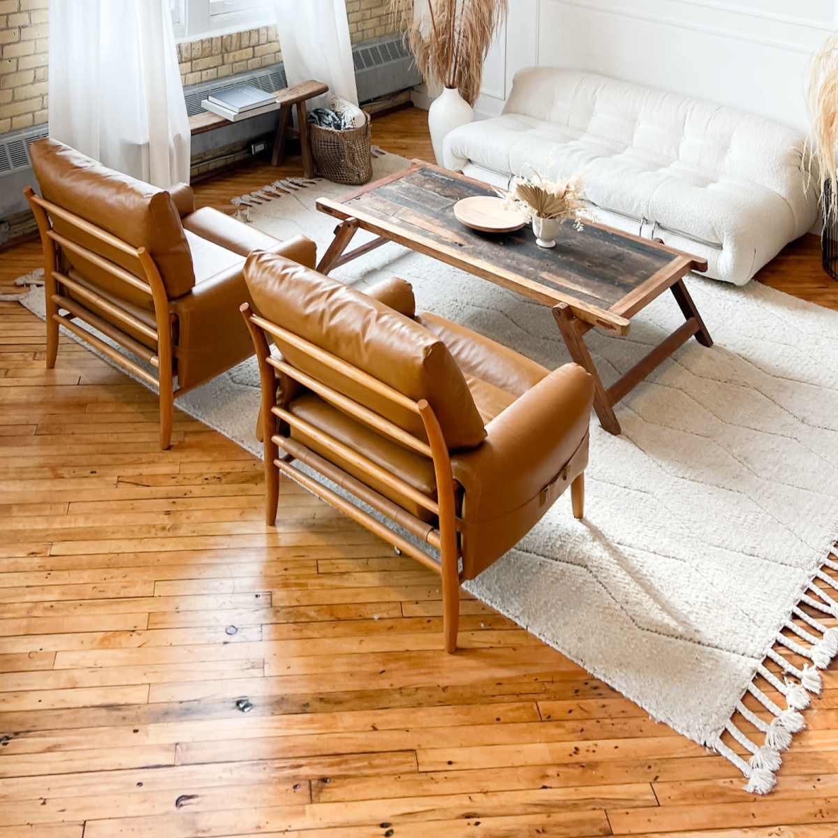 A modern living room features two brown leather chairs, a wooden coffee table, and a white couch, all set on a light rug over hardwood flooring with large windows allowing natural light.
