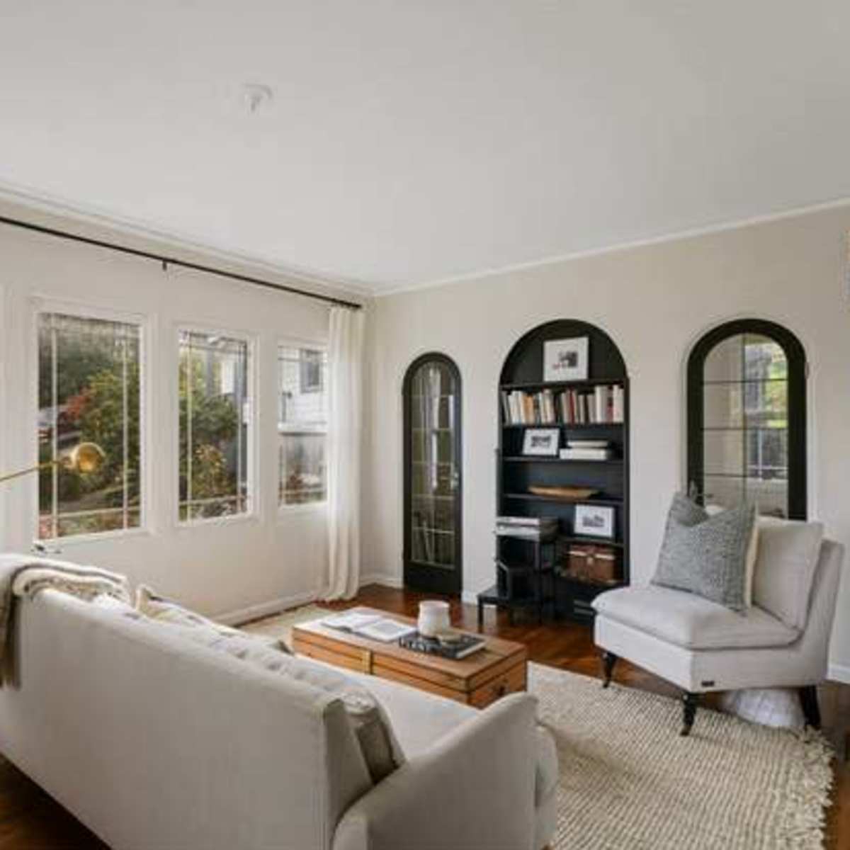 A cozy living room features a white sofa, a wooden coffee table, and large windows allowing natural light, with bookshelves and decorative arches in the background.