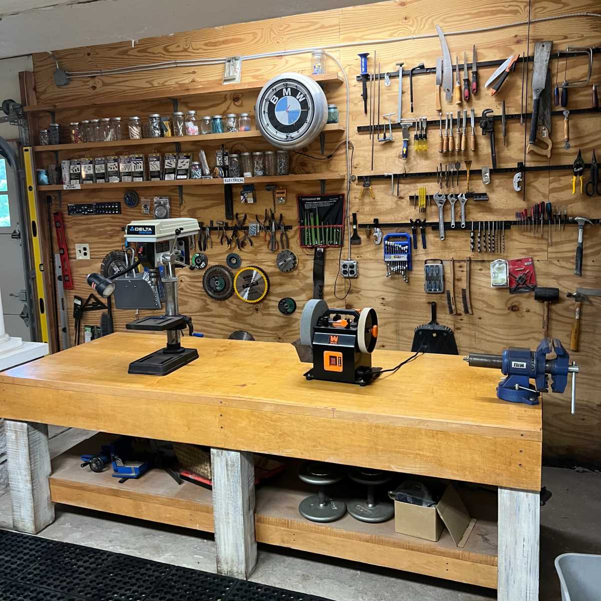 A well-organized workshop with a wooden workbench, various tools mounted on the wall, and equipment like a drill press, grinder, and vise displayed prominently.