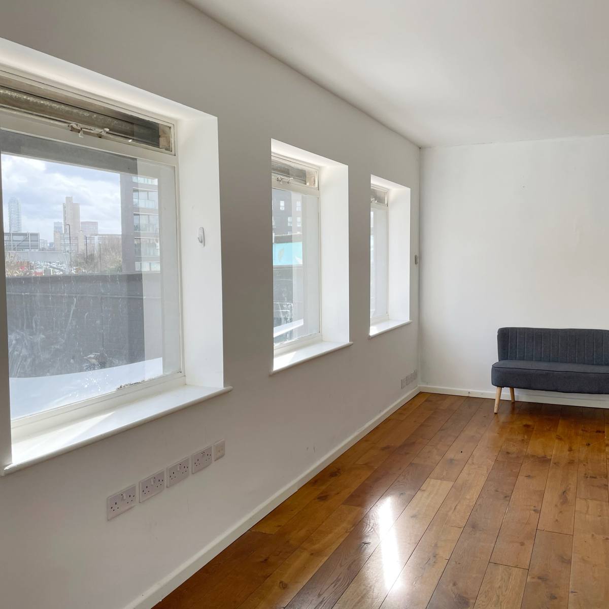A room with three windows and a gray couch against a white wall, featuring hardwood flooring.