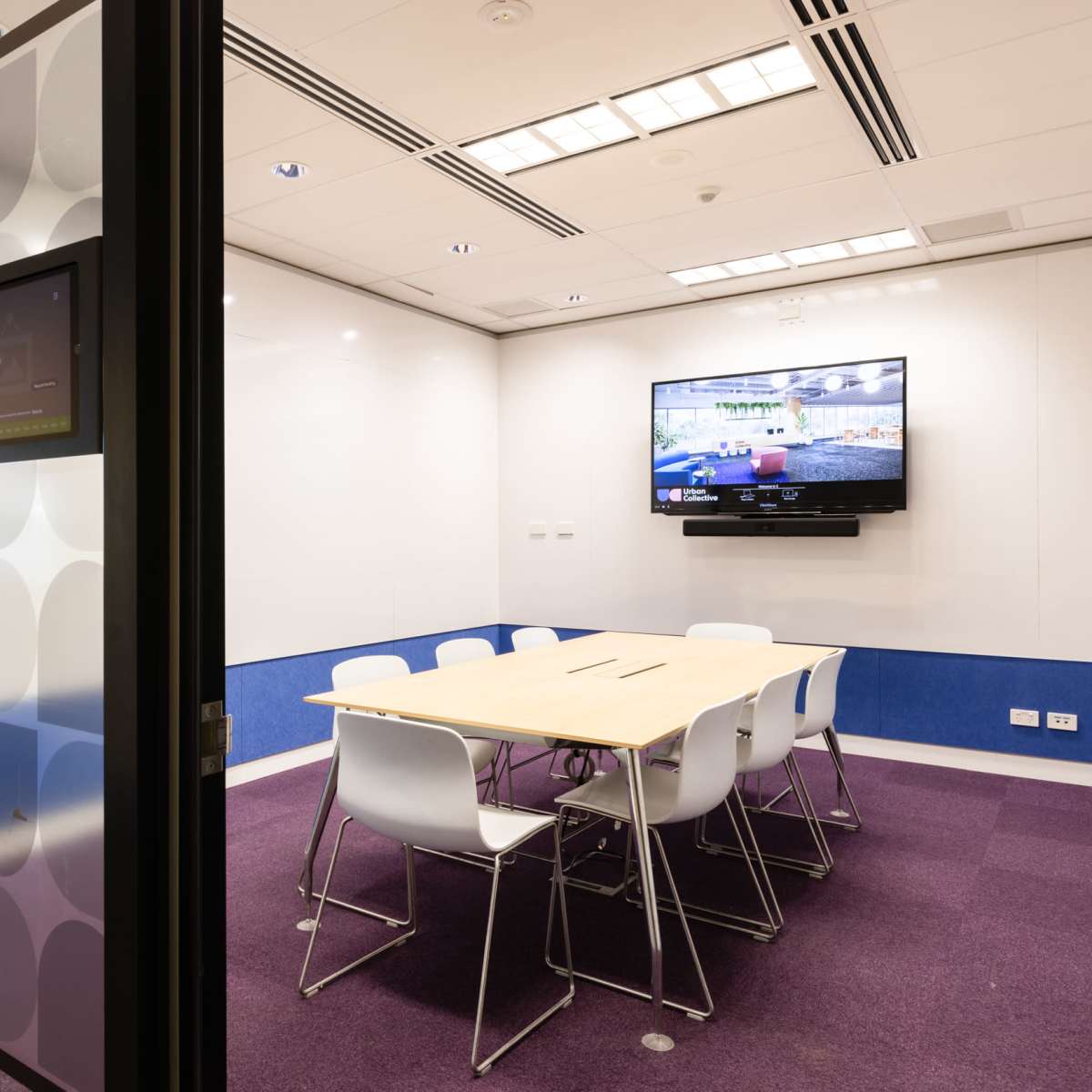 A conference room features a large table surrounded by white chairs, with a television mounted on the wall and a touchscreen control panel nearby.