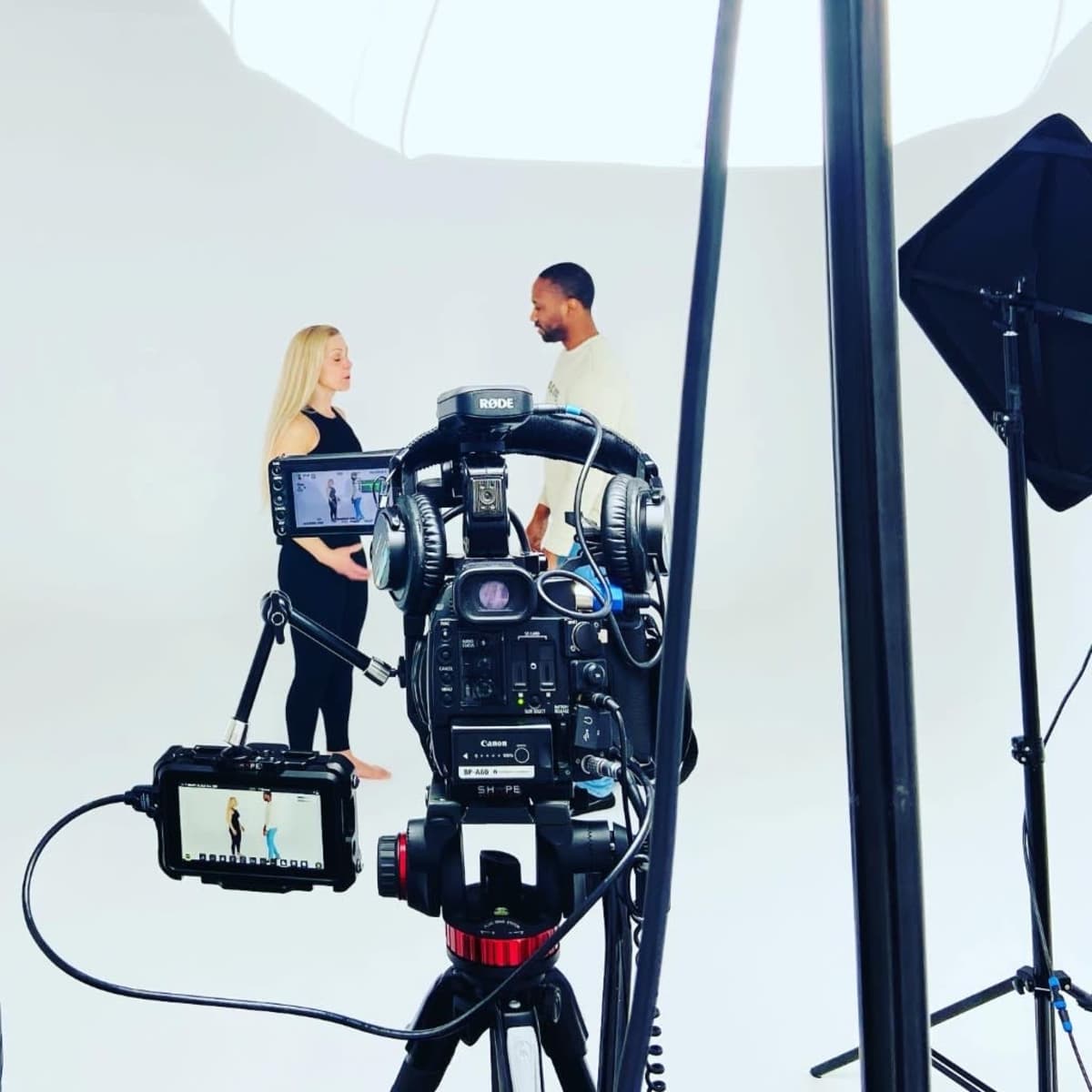 A camera set up on a tripod captures two individuals engaged in conversation in a well-lit studio against a plain white backdrop.