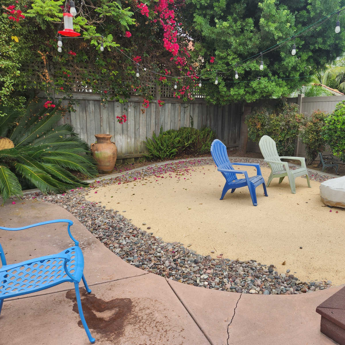 A small outdoor patio area with two colorful chairs and a covered fire pit surrounded by rocks and greenery.