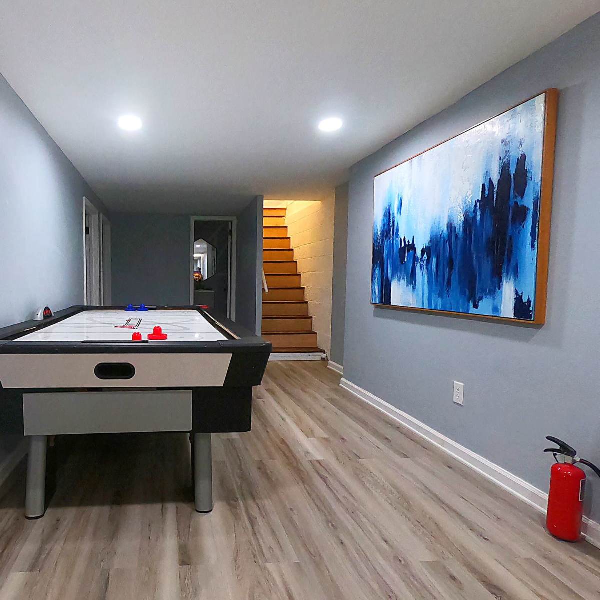 A game room features an air hockey table, a large abstract painting on the wall, and a staircase in the background.
