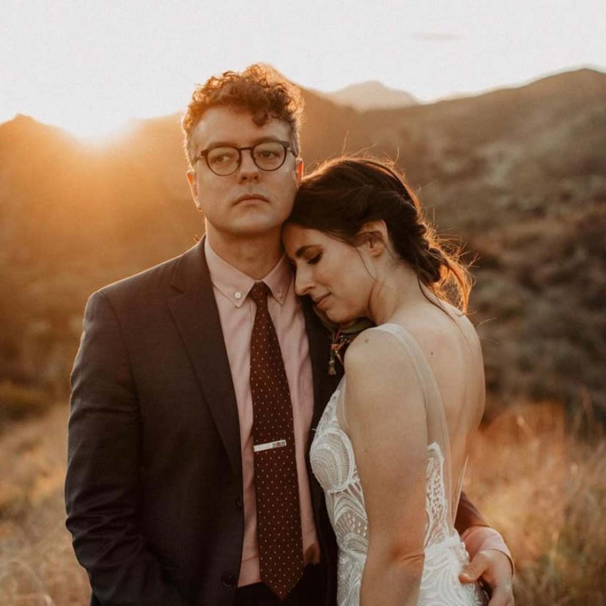 A man in a suit stands with a woman in a wedding dress, as they embrace in a sunlit, mountainous landscape.