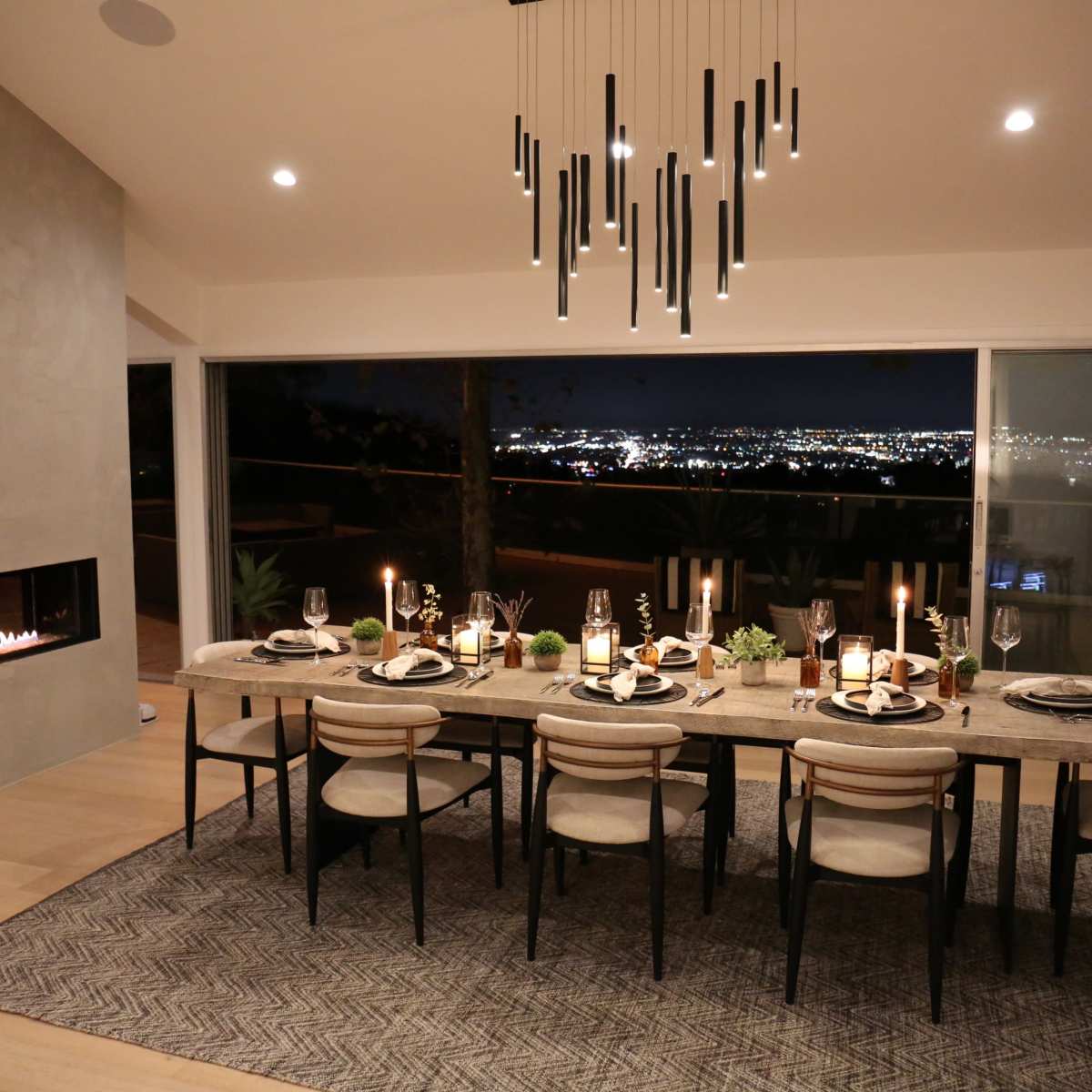 A modern dining room with a long table set for dinner, featuring a large window overlooking a nighttime city skyline and a sleek fireplace on the wall.