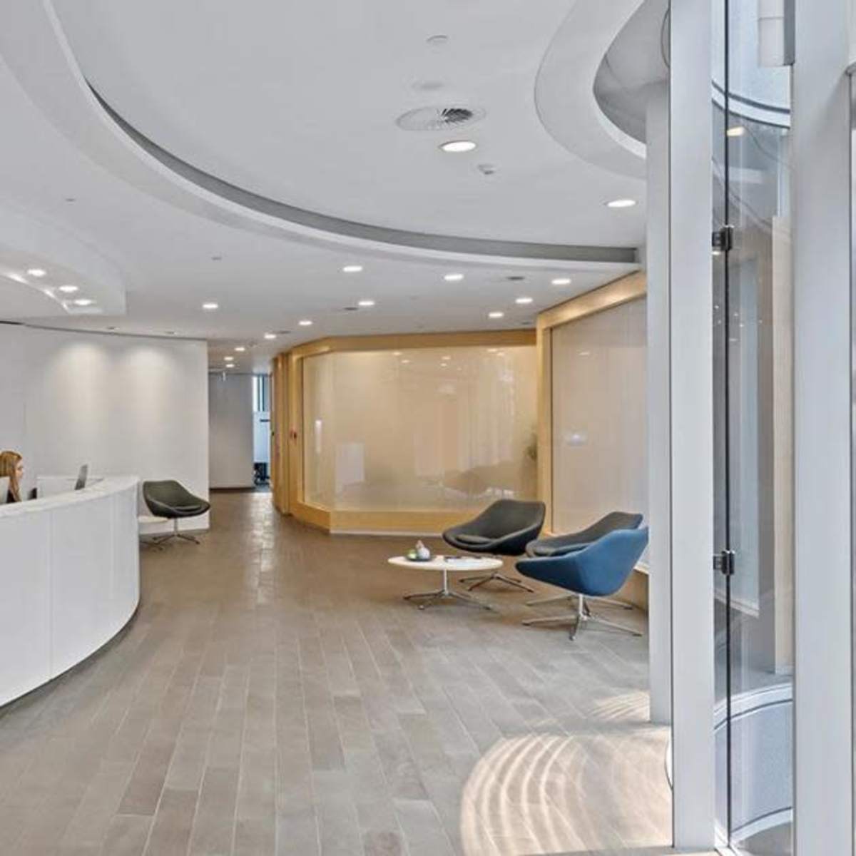 A modern reception area features a curved white desk with two staff members, surrounded by minimalistic furniture and a large window letting in natural light.
