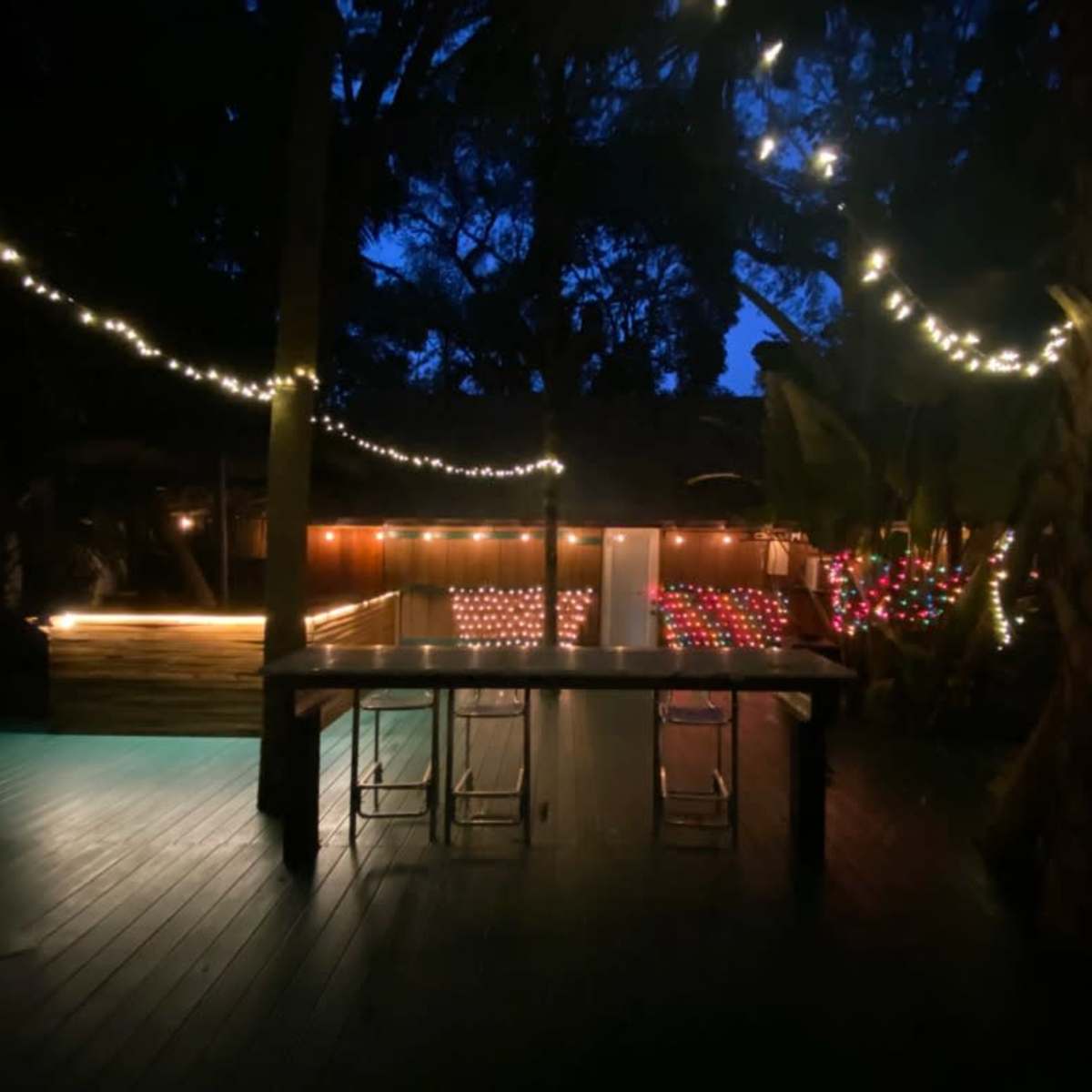 The scene features an outdoor area illuminated by string lights at dusk, with a wooden deck and a seating arrangement.