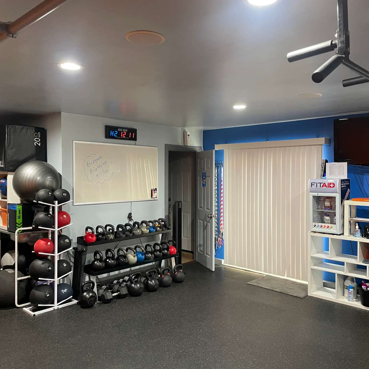 A neatly organized home gym with a rack of kettlebells, exercise balls, and a wall-mounted television in a well-lit room.