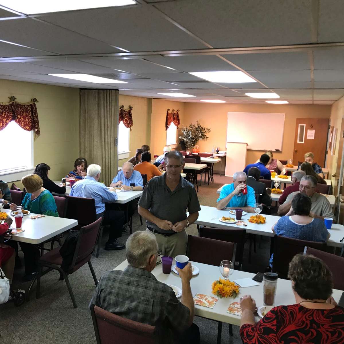 A group of people sits at tables in a community room, engaged in conversation and sharing a meal.