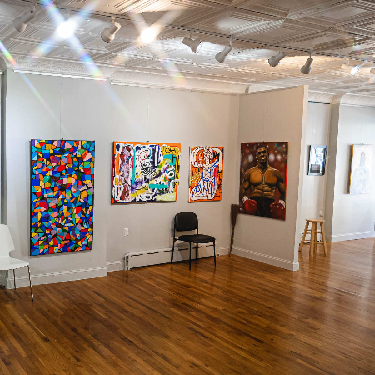 An art gallery with a wooden floor displaying several colorful paintings on the walls, along with a few chairs and a small counter in the corner.