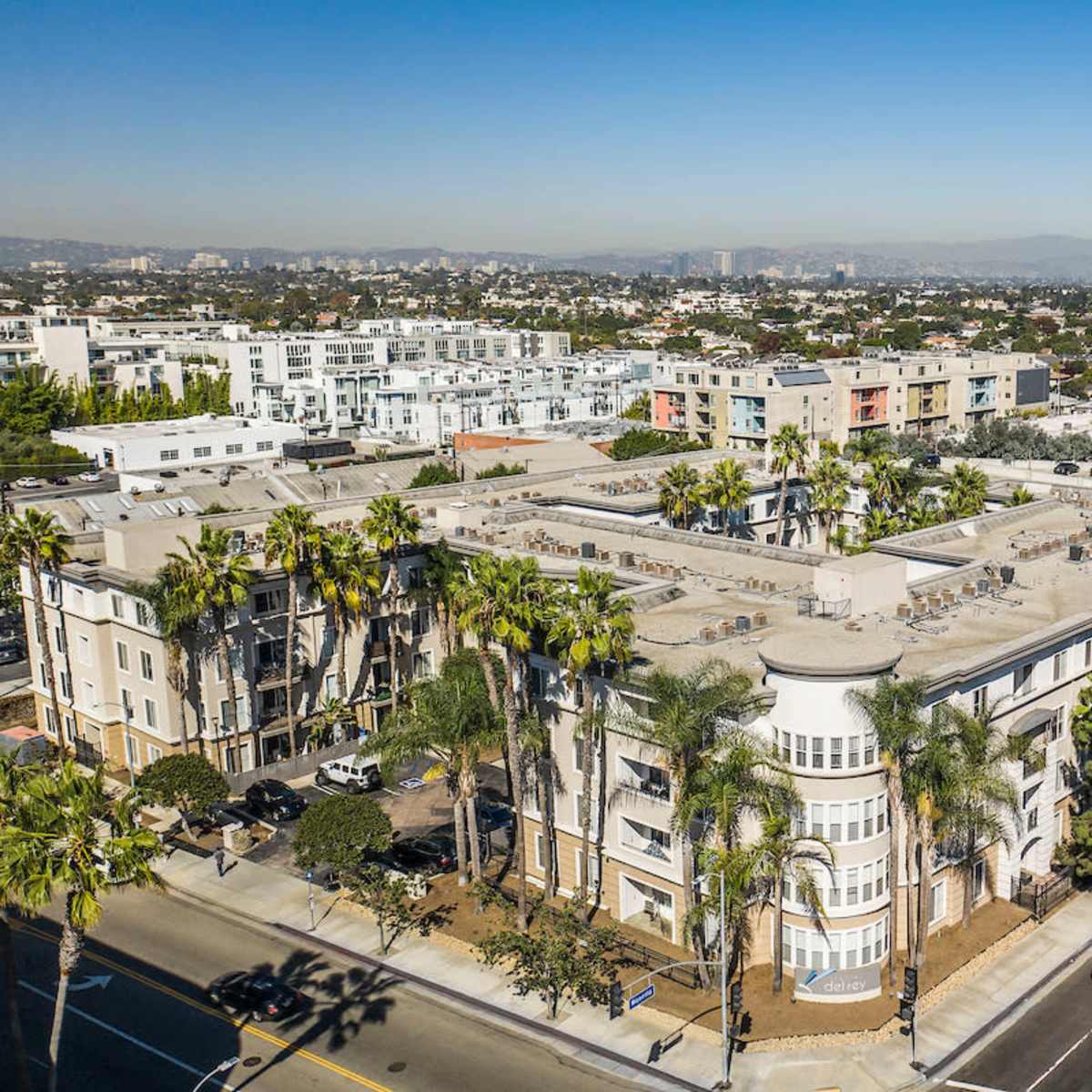 An aerial view shows a cluster of residential buildings surrounded by palm trees and a clear sky.