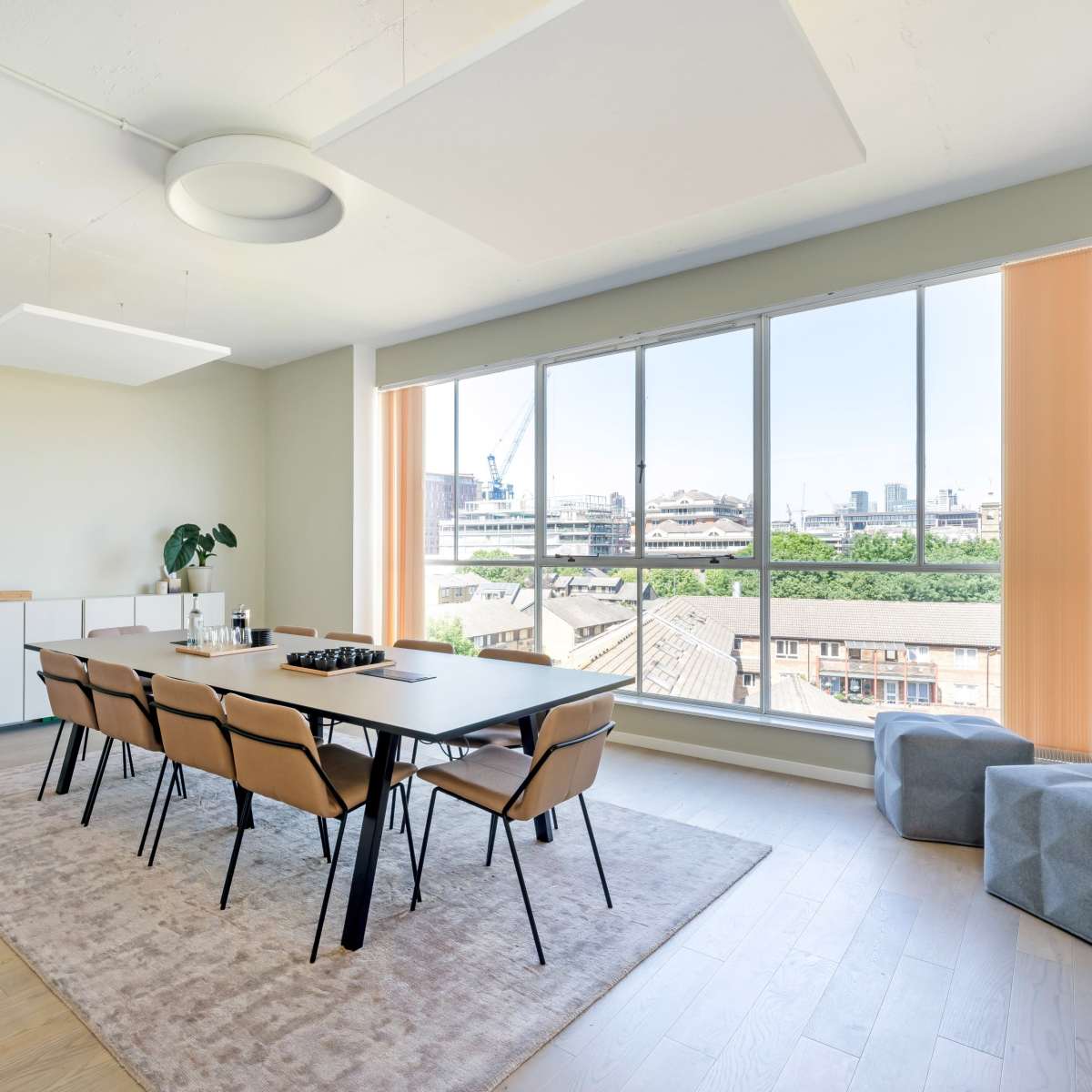 A modern dining area with a long table and chairs, large windows offering a view of the city, and minimalistic furniture.
