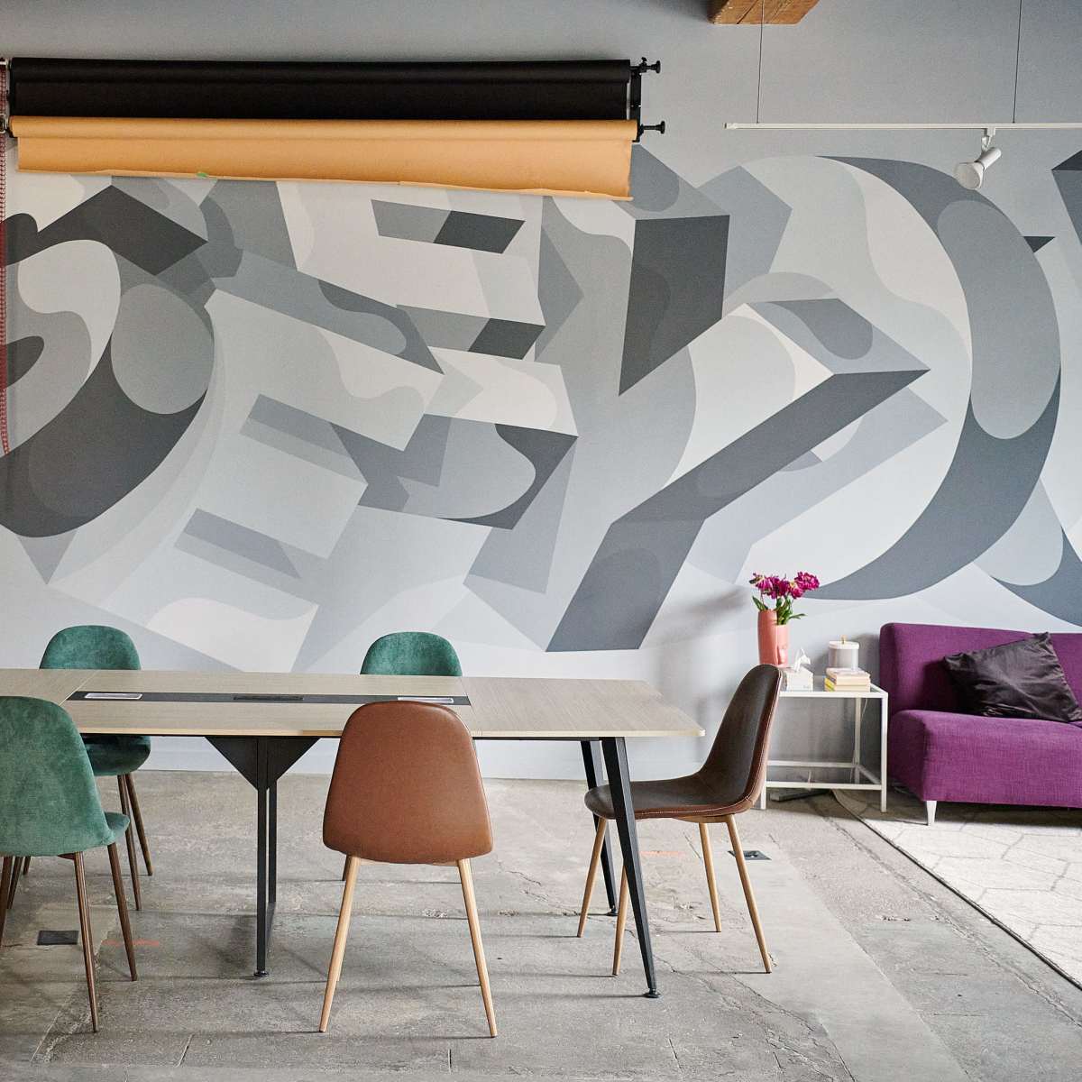 A modern meeting space features a wooden table surrounded by colored chairs, with a large mural in the background and a purple sofa to the side.