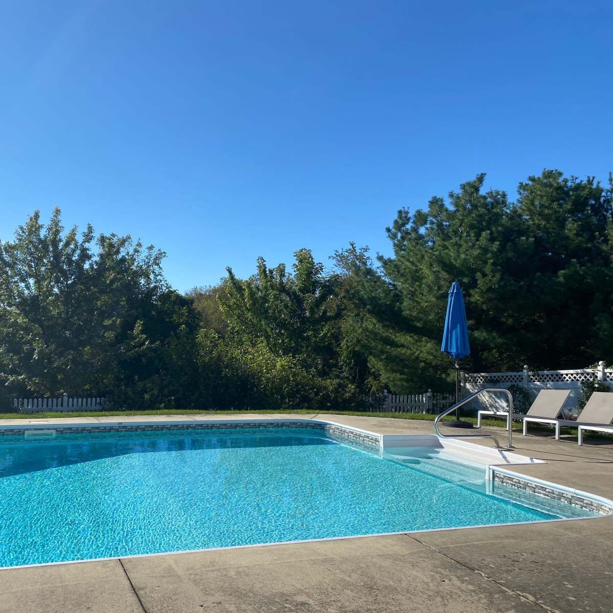 A clear swimming pool surrounded by green trees and lawn chairs under a blue umbrella.